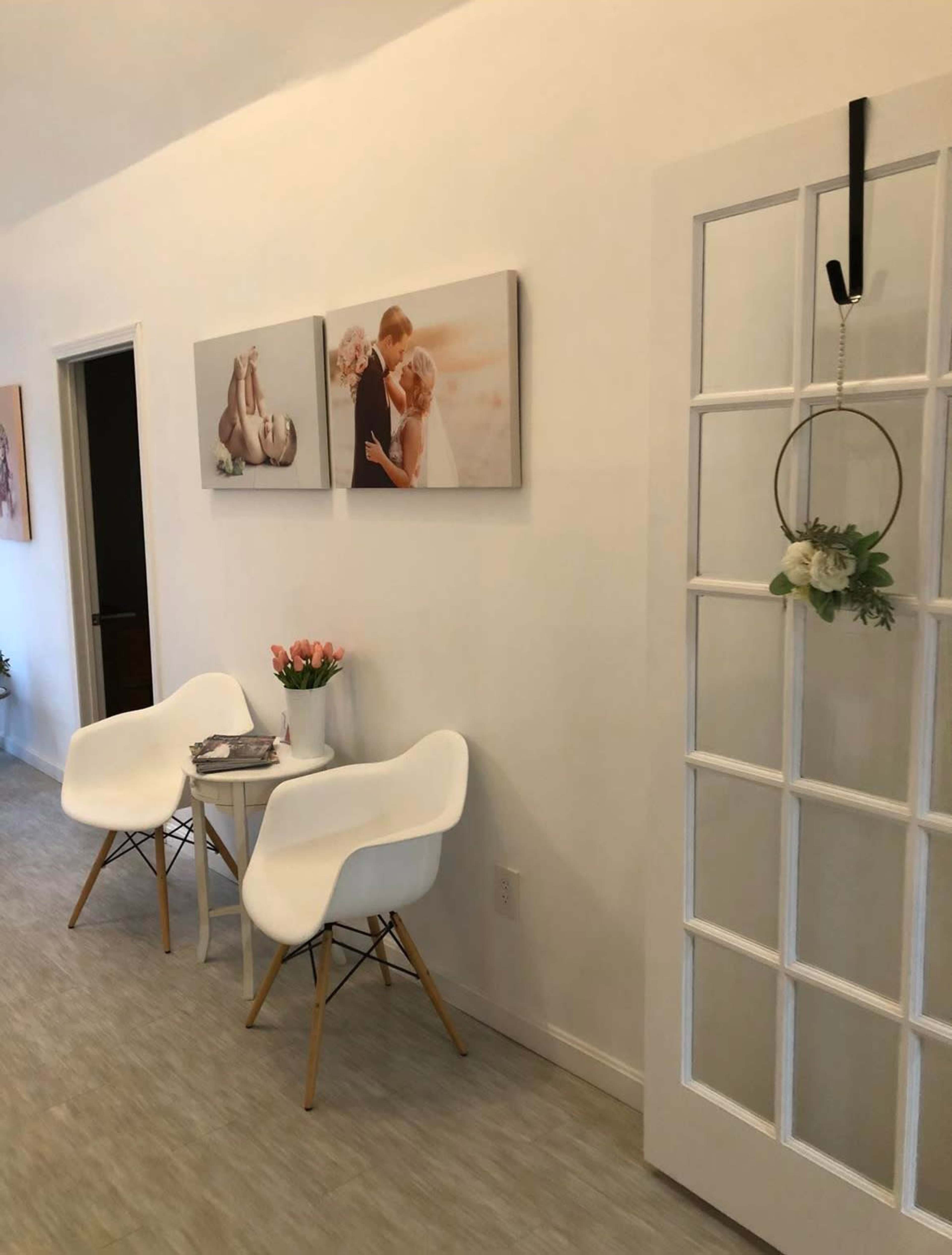 A bright, minimalist hallway featuring two white chairs, a small table with a flower vase, framed pictures on the wall, and a glass-paneled door.