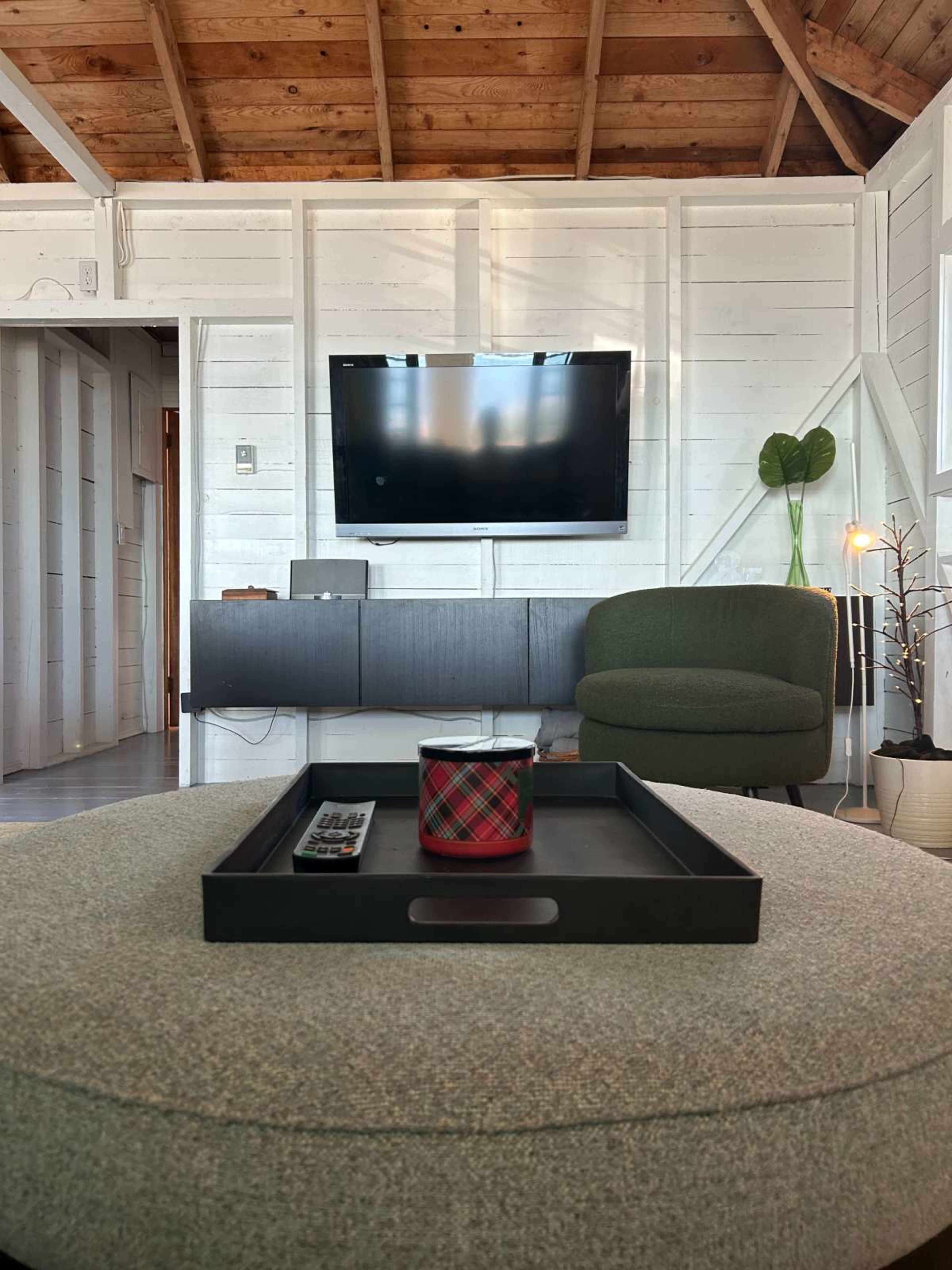 The image shows a modern living room featuring a television mounted on a white wooden wall, with a green armchair, a coffee table with a tray and candle, and an indoor plant in the corner.