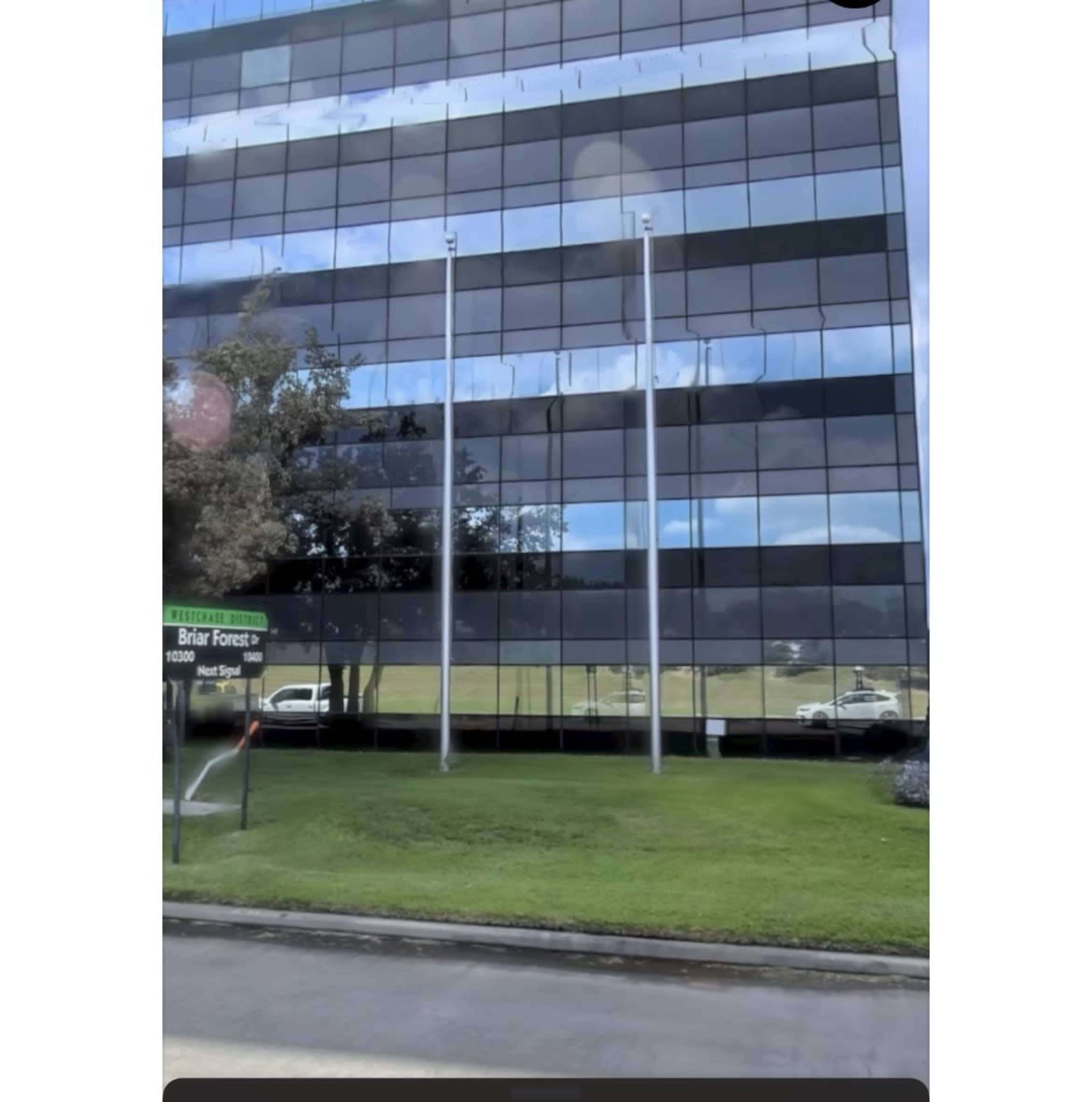 The image shows a modern glass office building reflecting clouds and a grassy area with flagpoles in front.