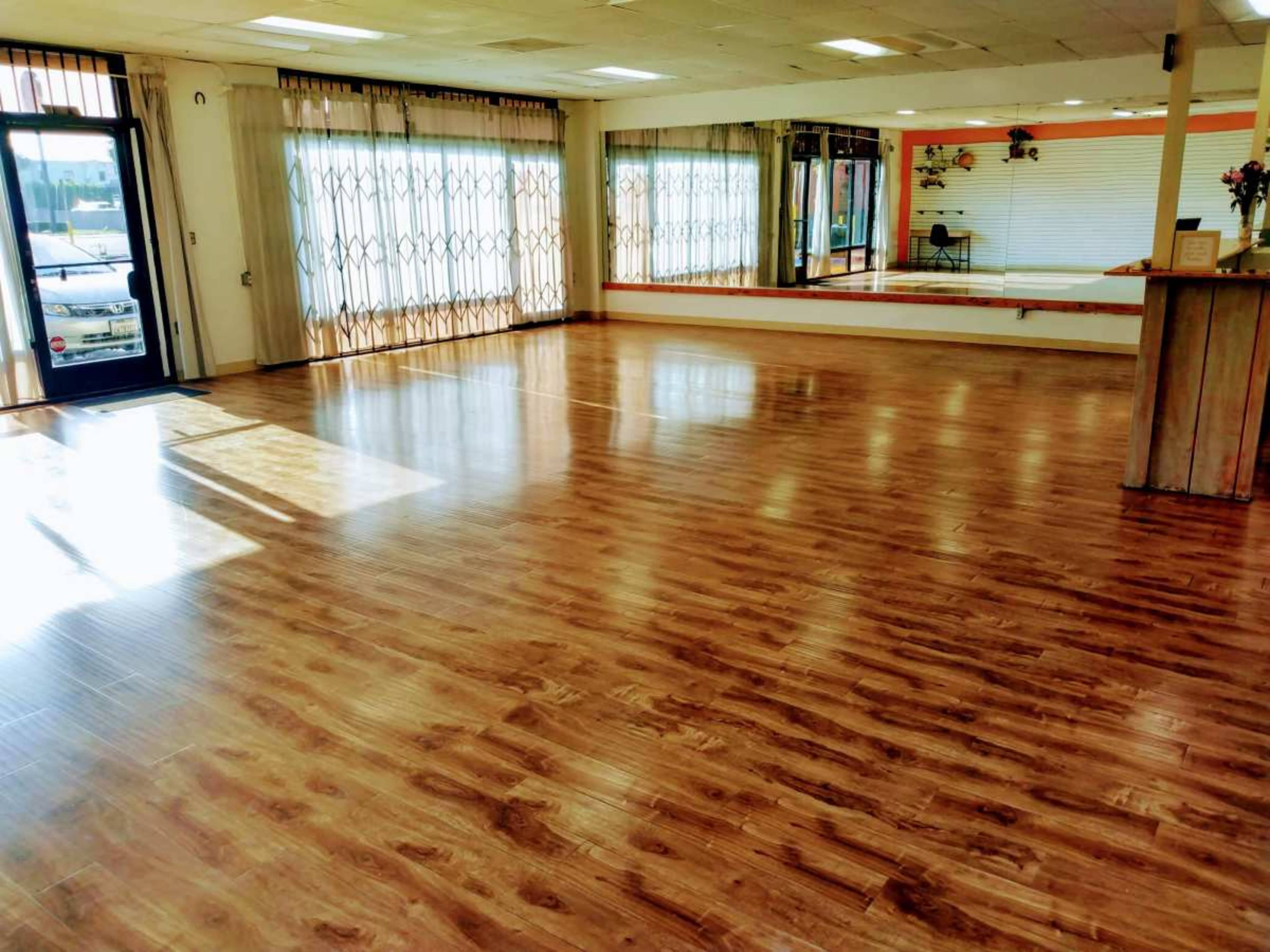 A spacious room with polished wooden flooring, large windows allowing natural light, and a mirrored wall.