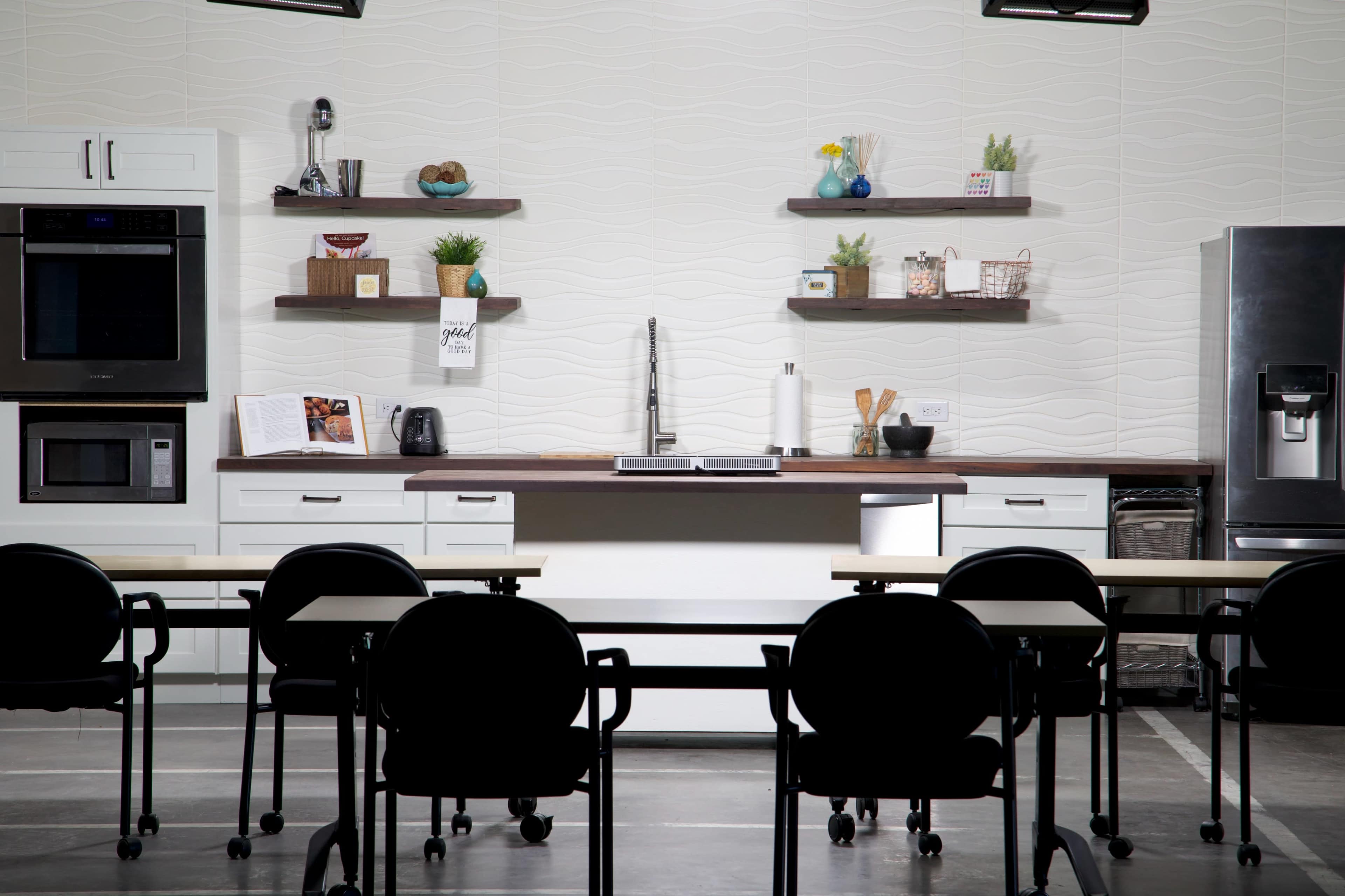 A modern kitchen setup with a cooking area, open shelving, and several appliances, positioned in front of a row of black chairs and tables.
