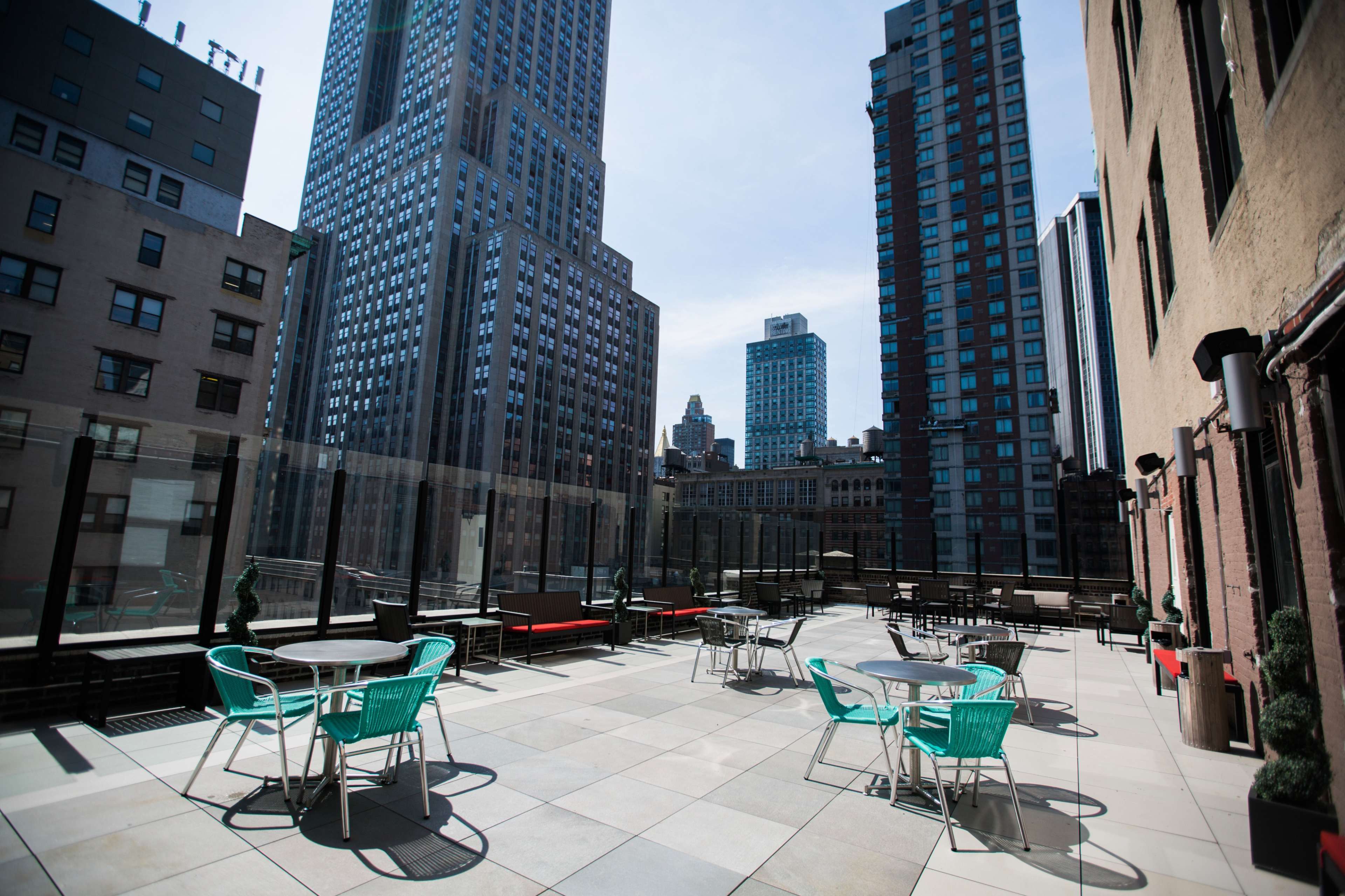 The image shows a rooftop terrace in an urban setting, featuring several round tables and chairs, surrounded by tall skyscrapers.