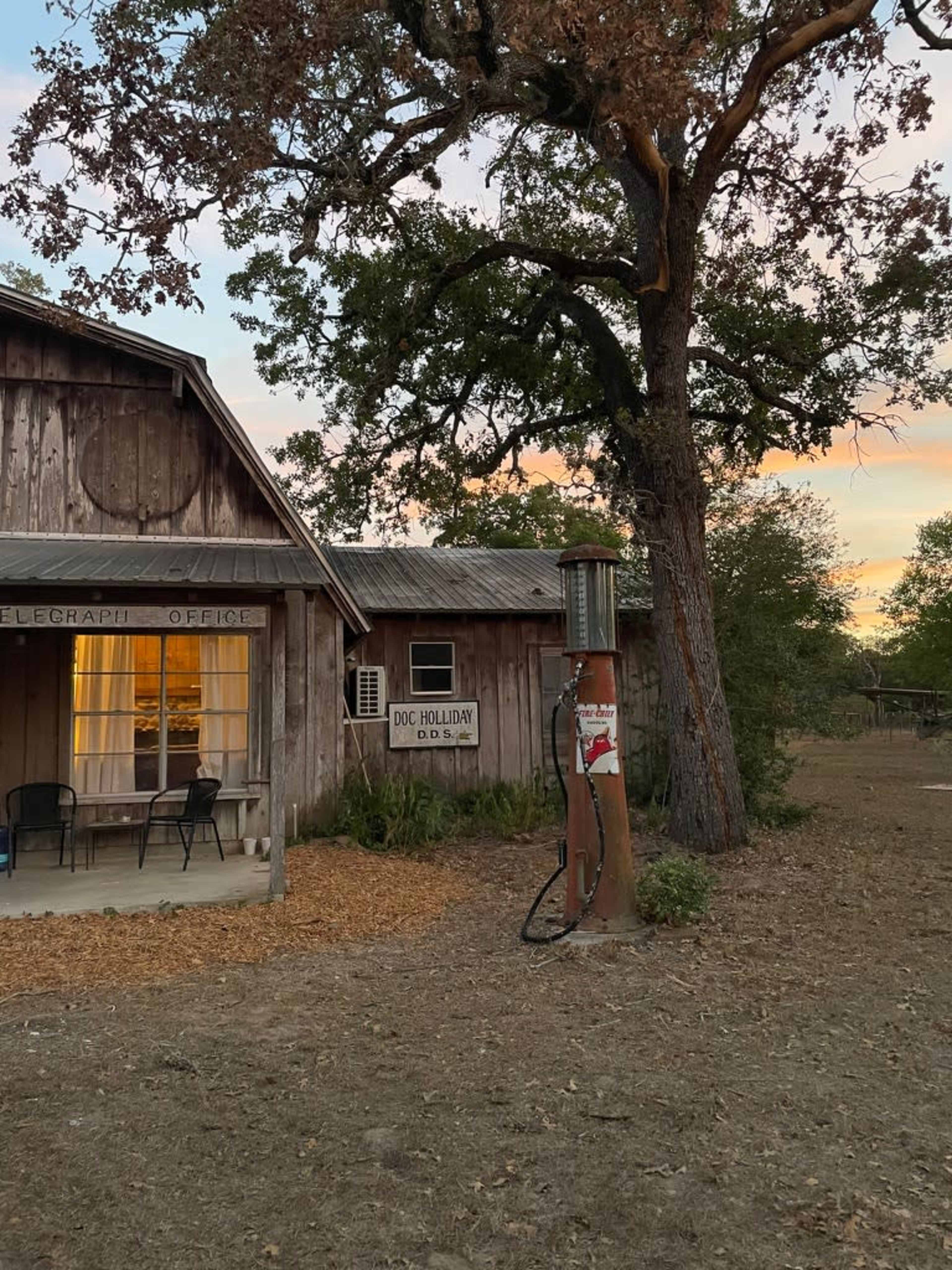 A weathered wooden building with a sign reading "Doc Holliday" and an old-fashioned gas pump in front, set against a sunset sky.
