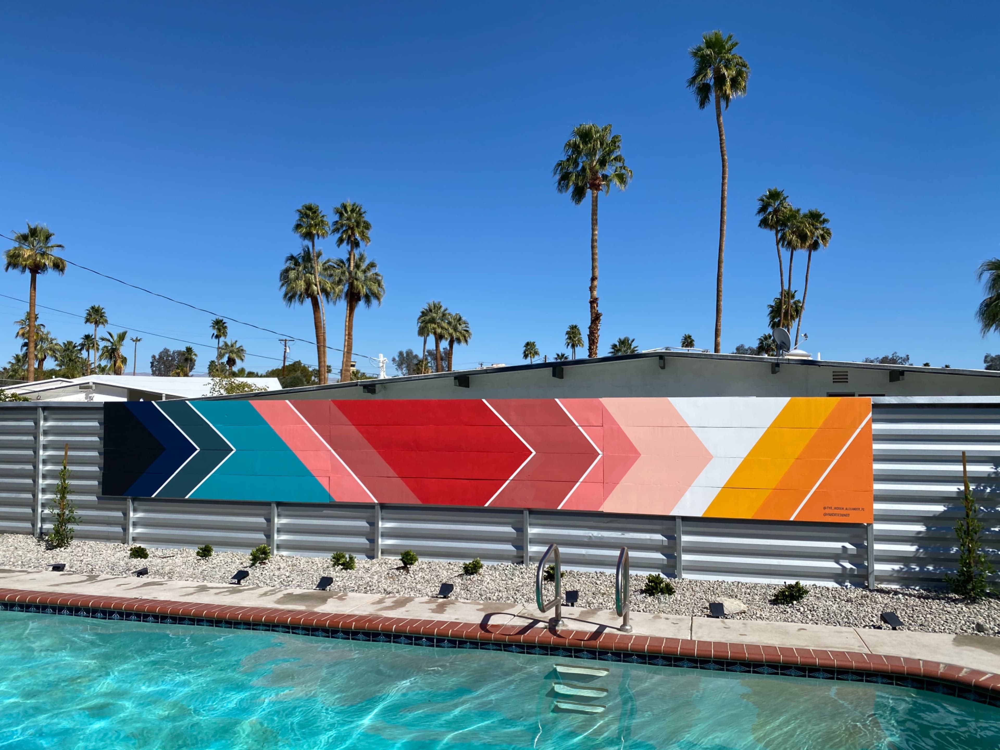 A colorful geometric mural featuring zigzag patterns is displayed on a wall next to a swimming pool, surrounded by palm trees under a clear blue sky.
