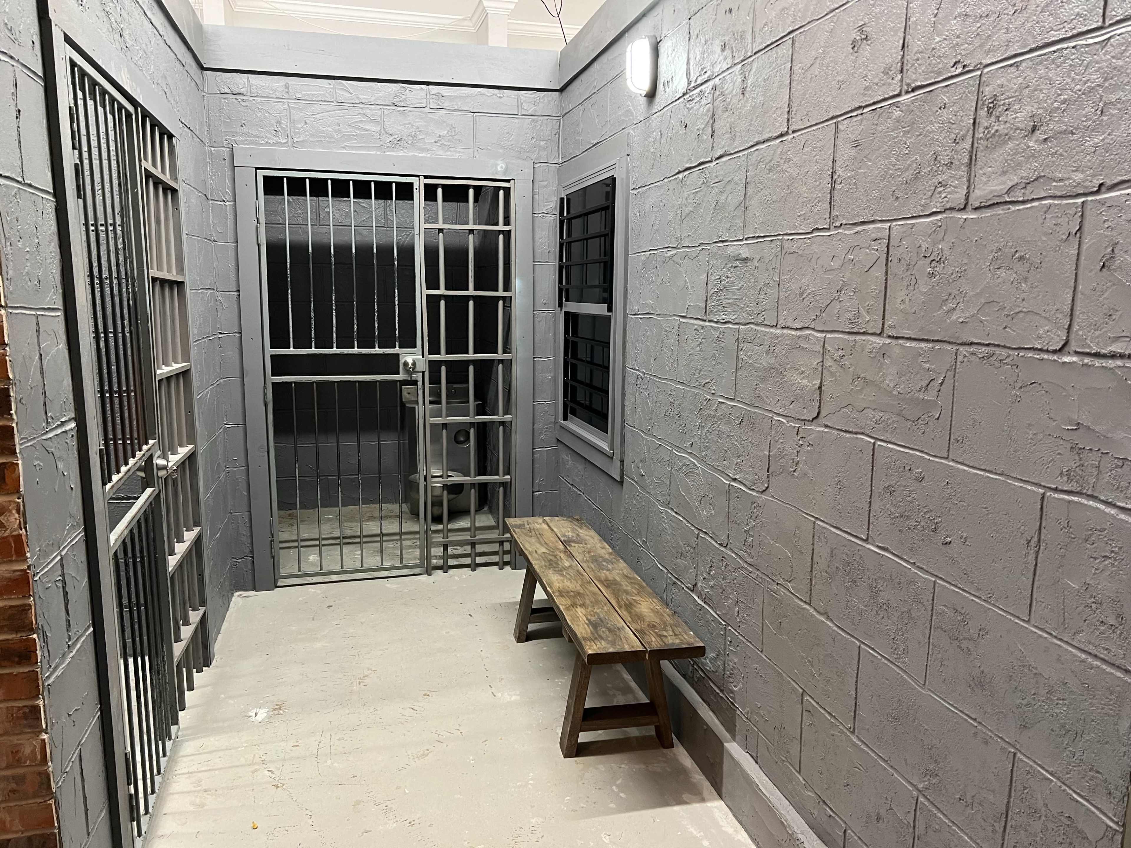 The image depicts a gray-walled hallway with prison-style bars, featuring a wooden bench and a locked metal gate.