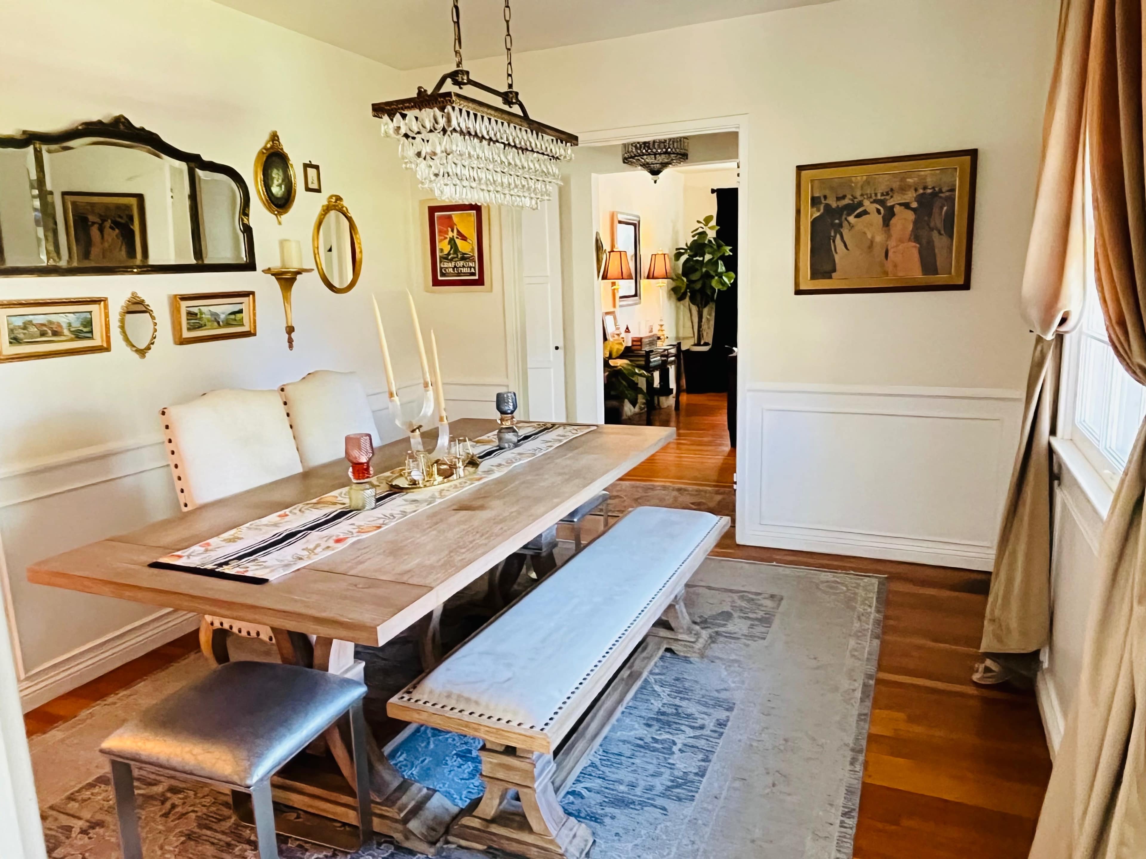 The image shows a dining room with a wooden table and bench, ornate chandelier, and decorative wall mirrors.