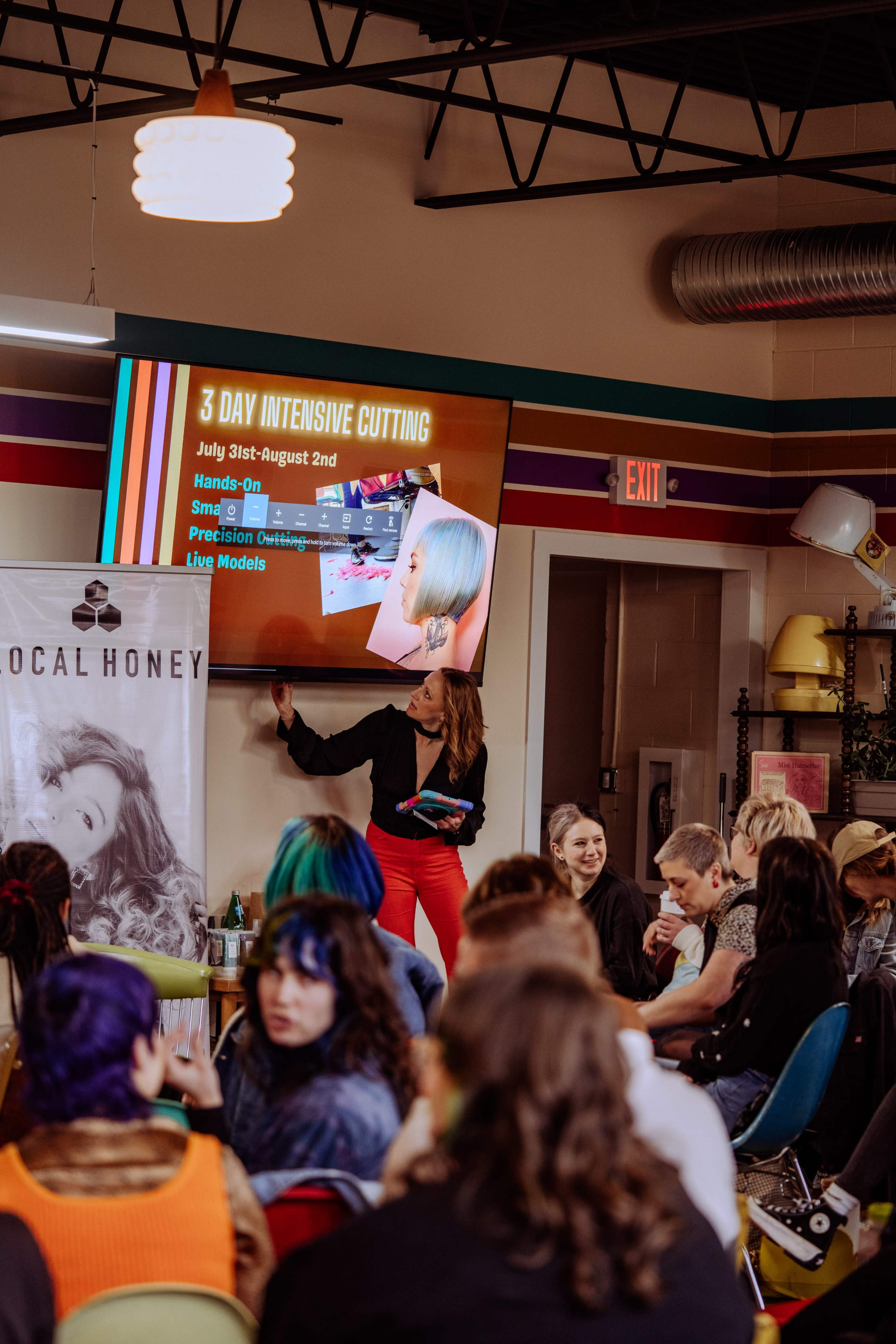 A woman stands in front of a screen displaying information about a three-day intensive cutting workshop, while a diverse group of people sits audience-style in a vibrant, colorful salon setting.