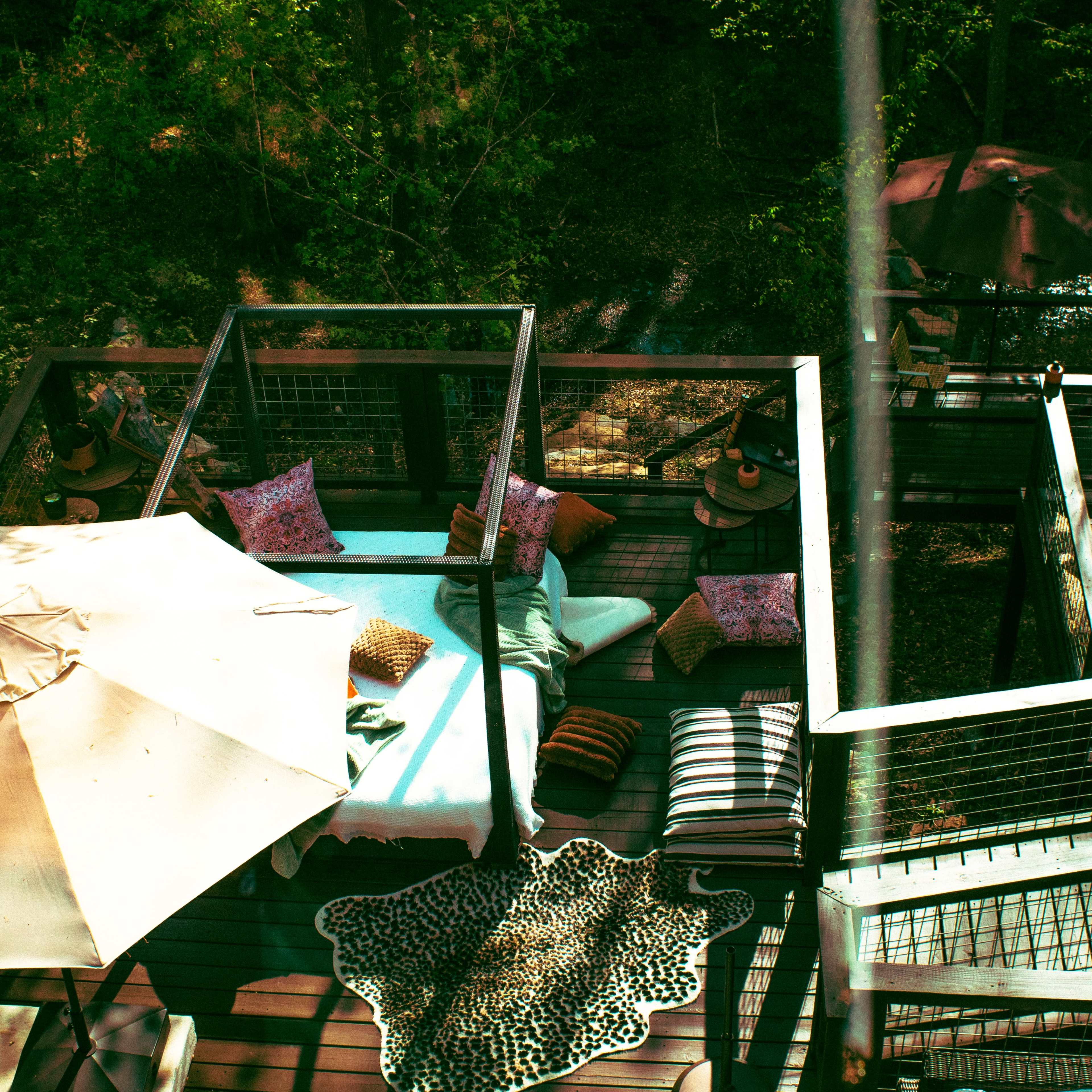 The image shows a wooden deck furnished with cushions, an umbrella, and a decorative rug, surrounded by trees.