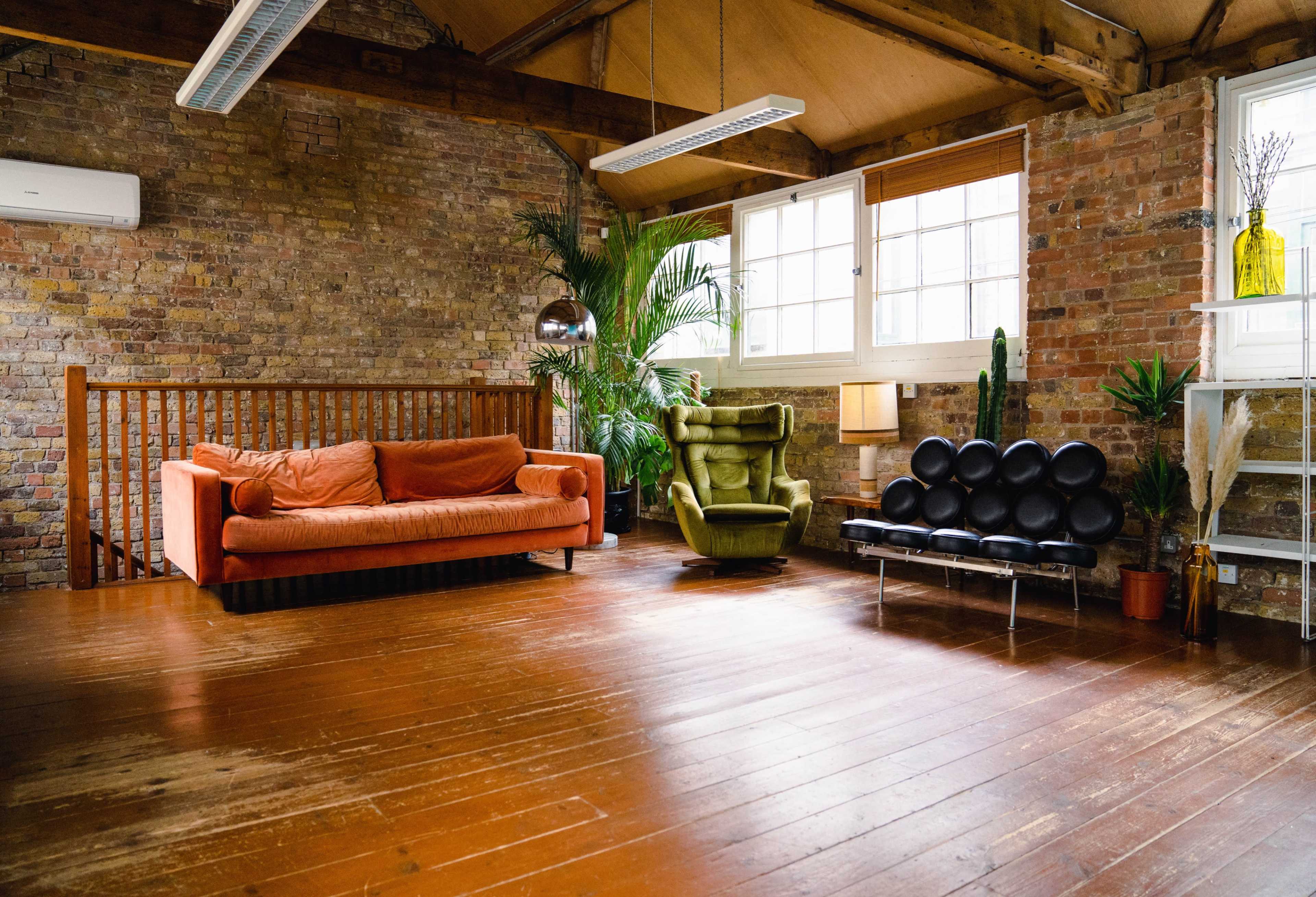 A spacious room with exposed brick walls, featuring an orange sofa, a green armchair, and a black modern seating arrangement, complemented by indoor plants and large windows.
