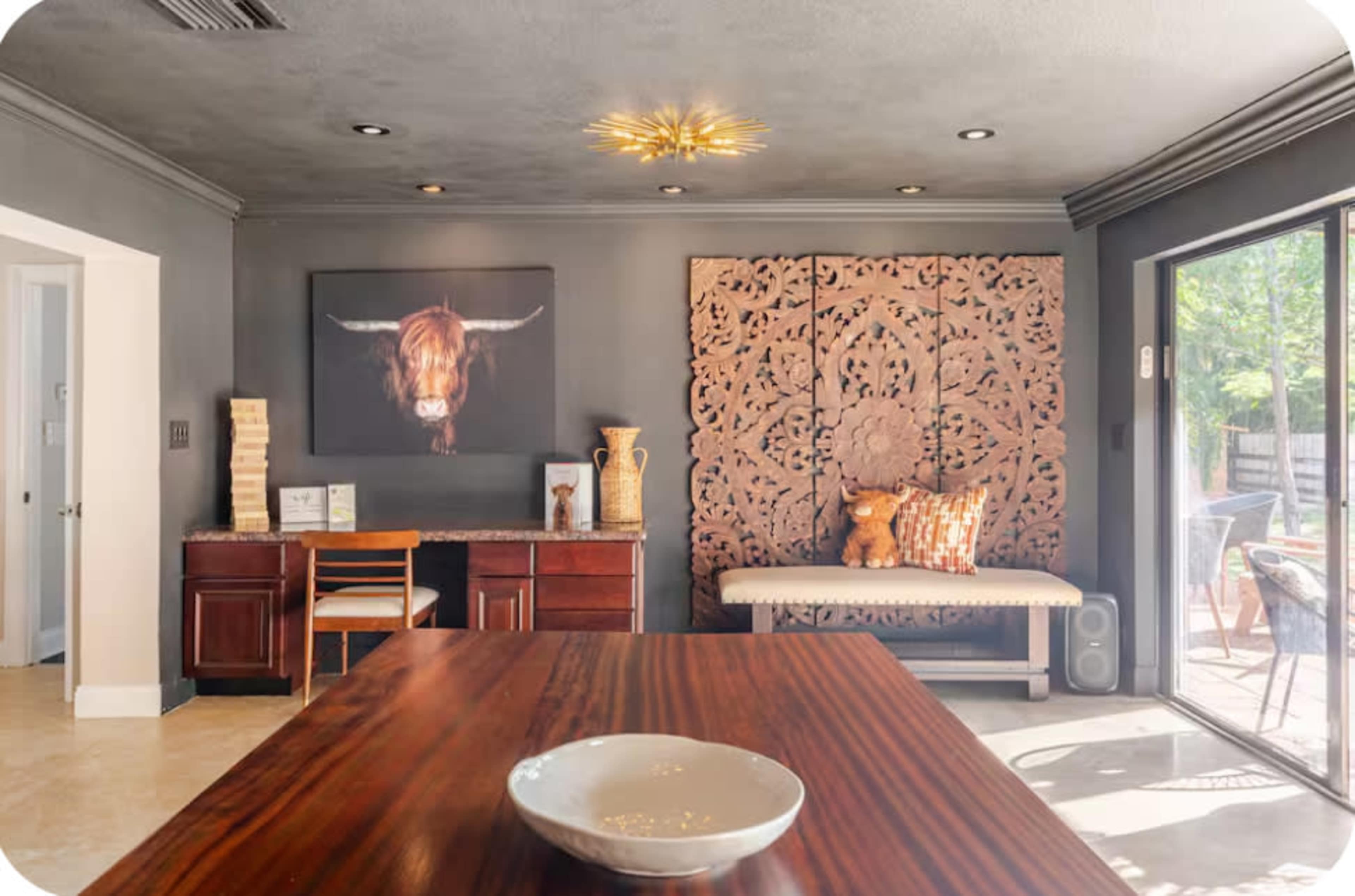 The image shows a modern living space with a wooden dining table in the foreground, a decorative wall panel, and a seating area featuring a sculpture and artwork of a bull's head.