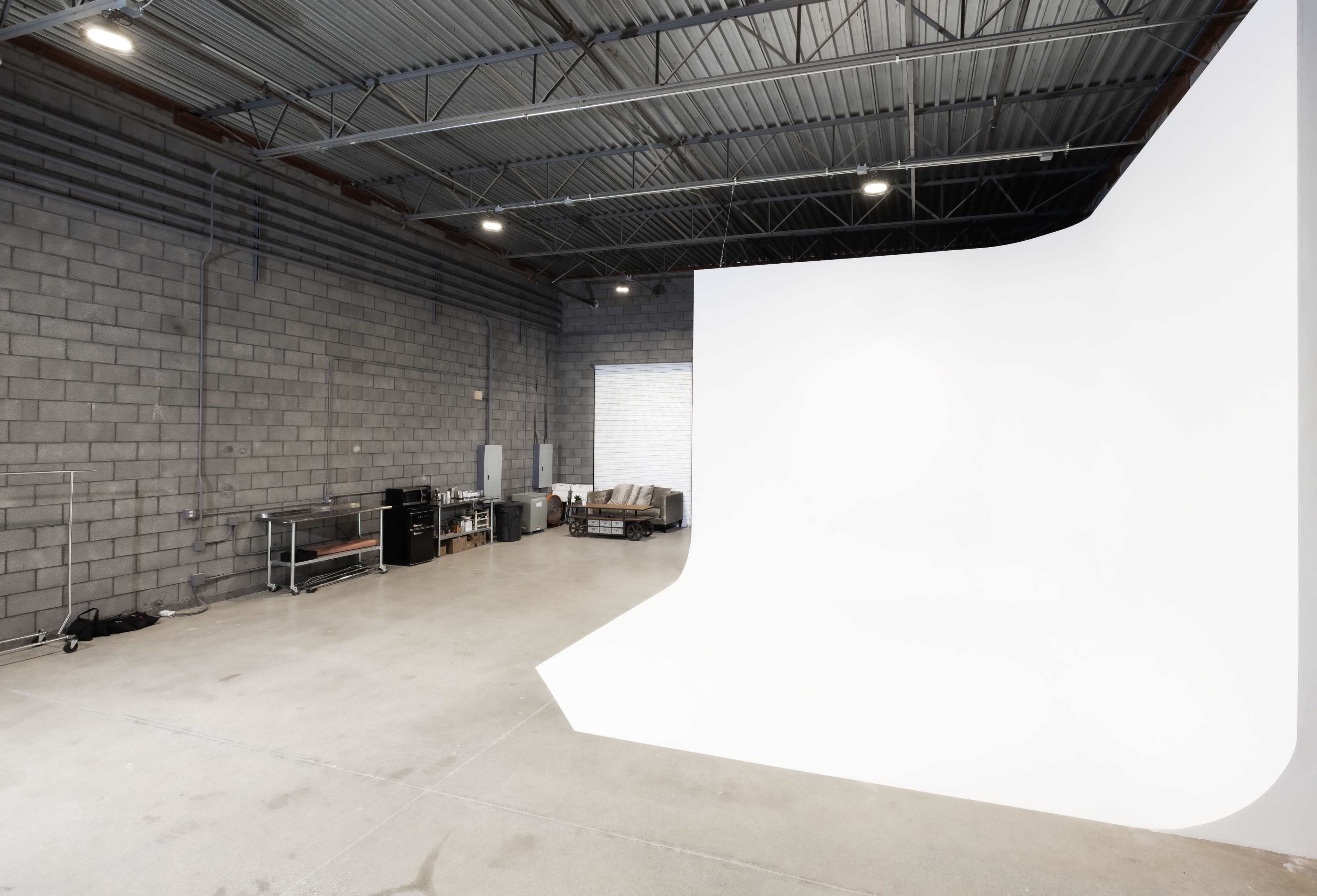 The image shows a spacious studio with a large white backdrop, industrial gray walls, and various furniture and equipment along one side.