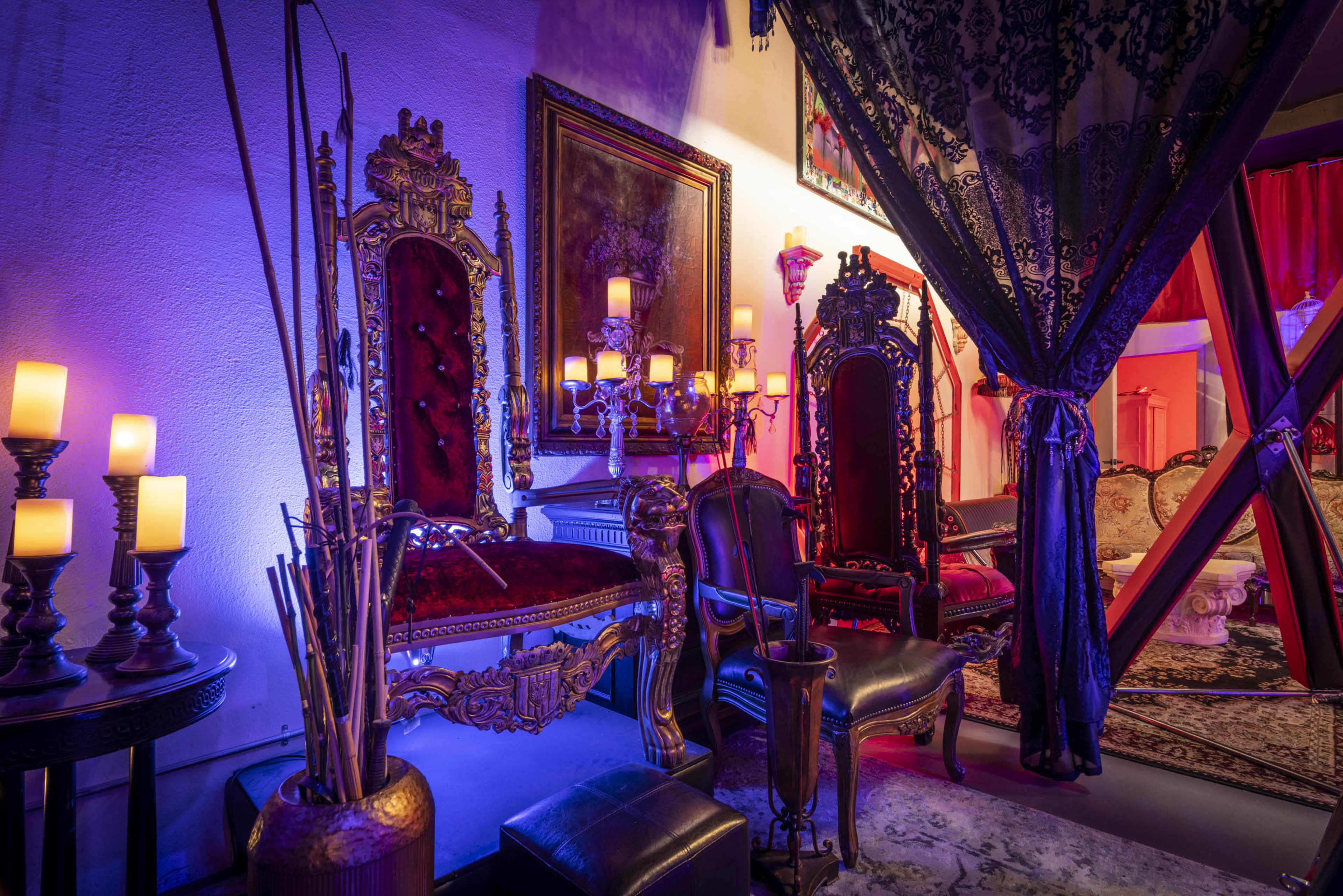 The image shows two ornate throne-like chairs beside a table with candles, set in a dimly lit room adorned with decorative fabrics and artwork.