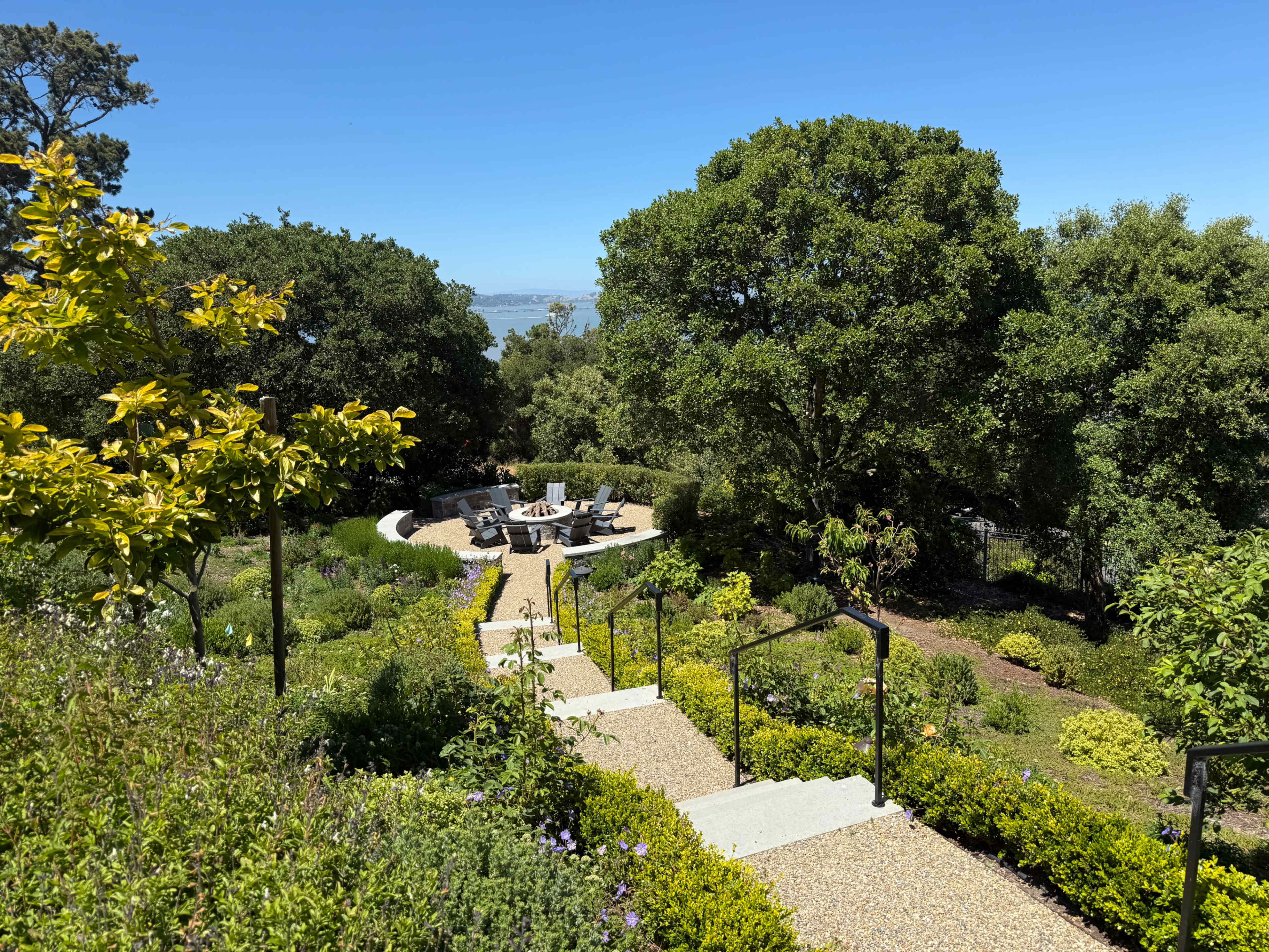 The image shows a landscaped garden with stone steps leading down to a circular seating area surrounded by trees and greenery.