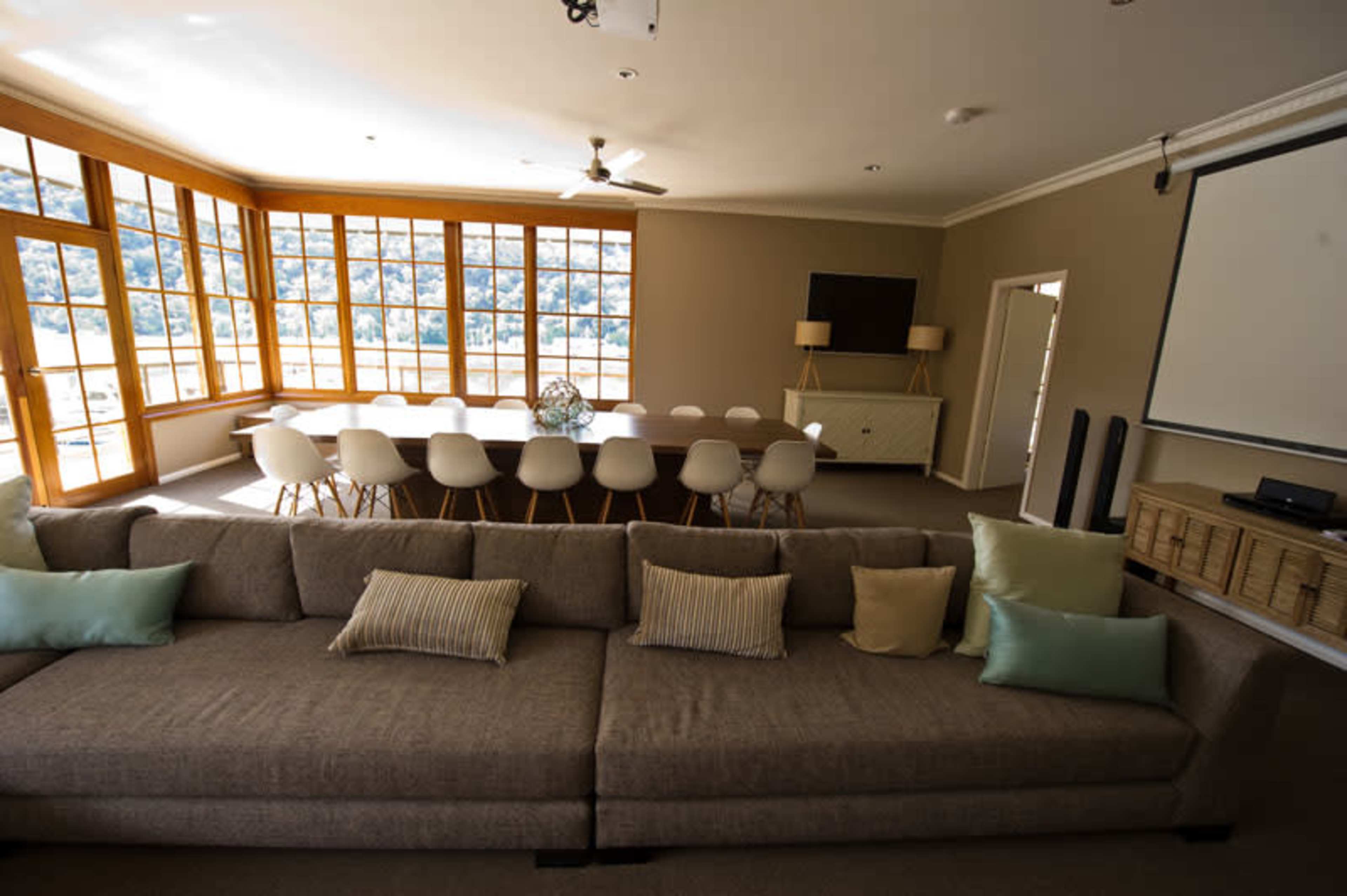 A spacious meeting room with a large table surrounded by chairs, large windows allowing natural light, and a comfortable couch with decorative pillows in the foreground.