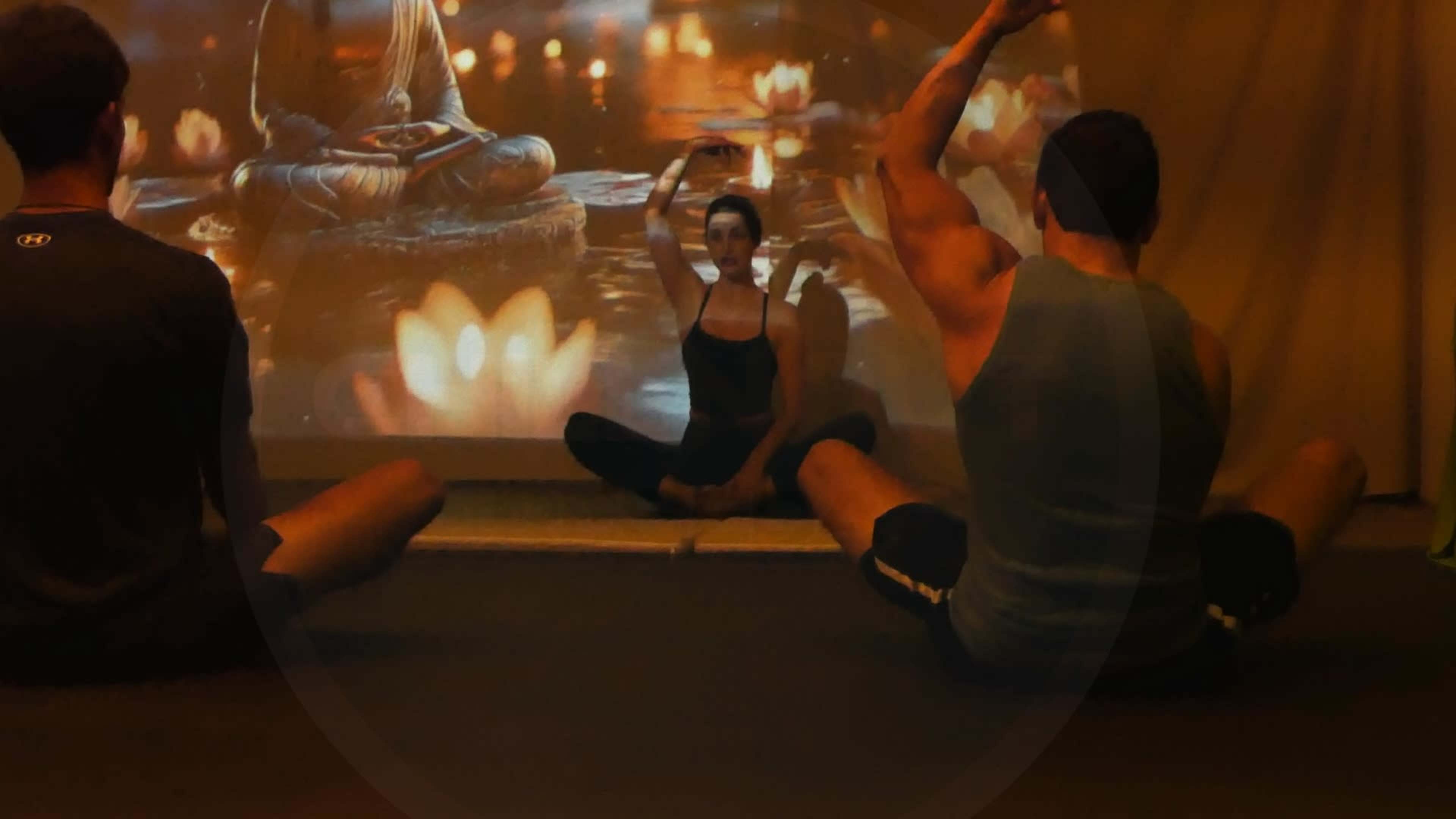 Three individuals practice yoga in a dimly lit room with a projection of a serene water scene featuring lotus flowers and a Buddha statue.