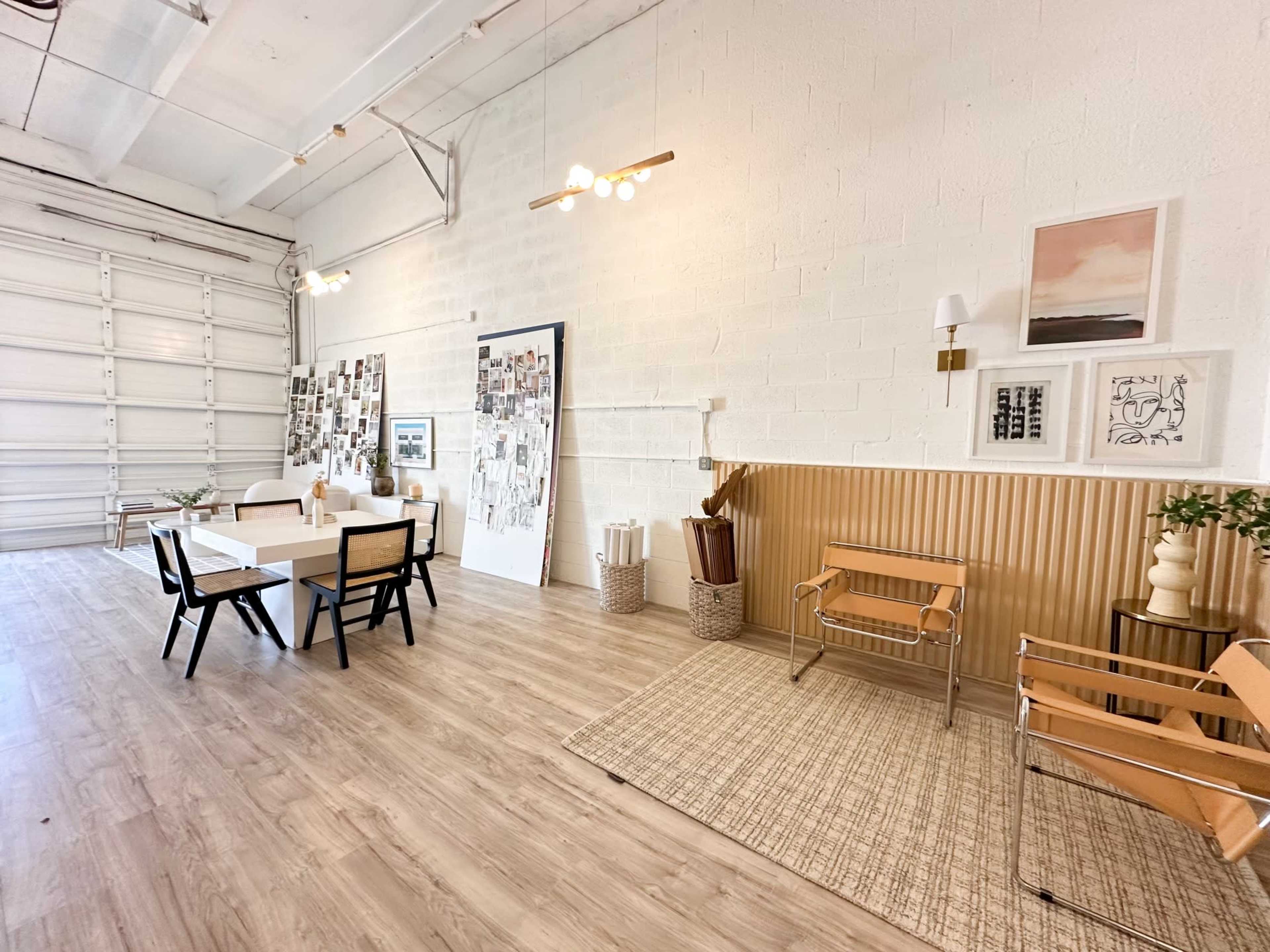 A spacious room with a large table and chairs, decorated with wall art and a gallery of photos, featuring light wood flooring and modern lighting fixtures.