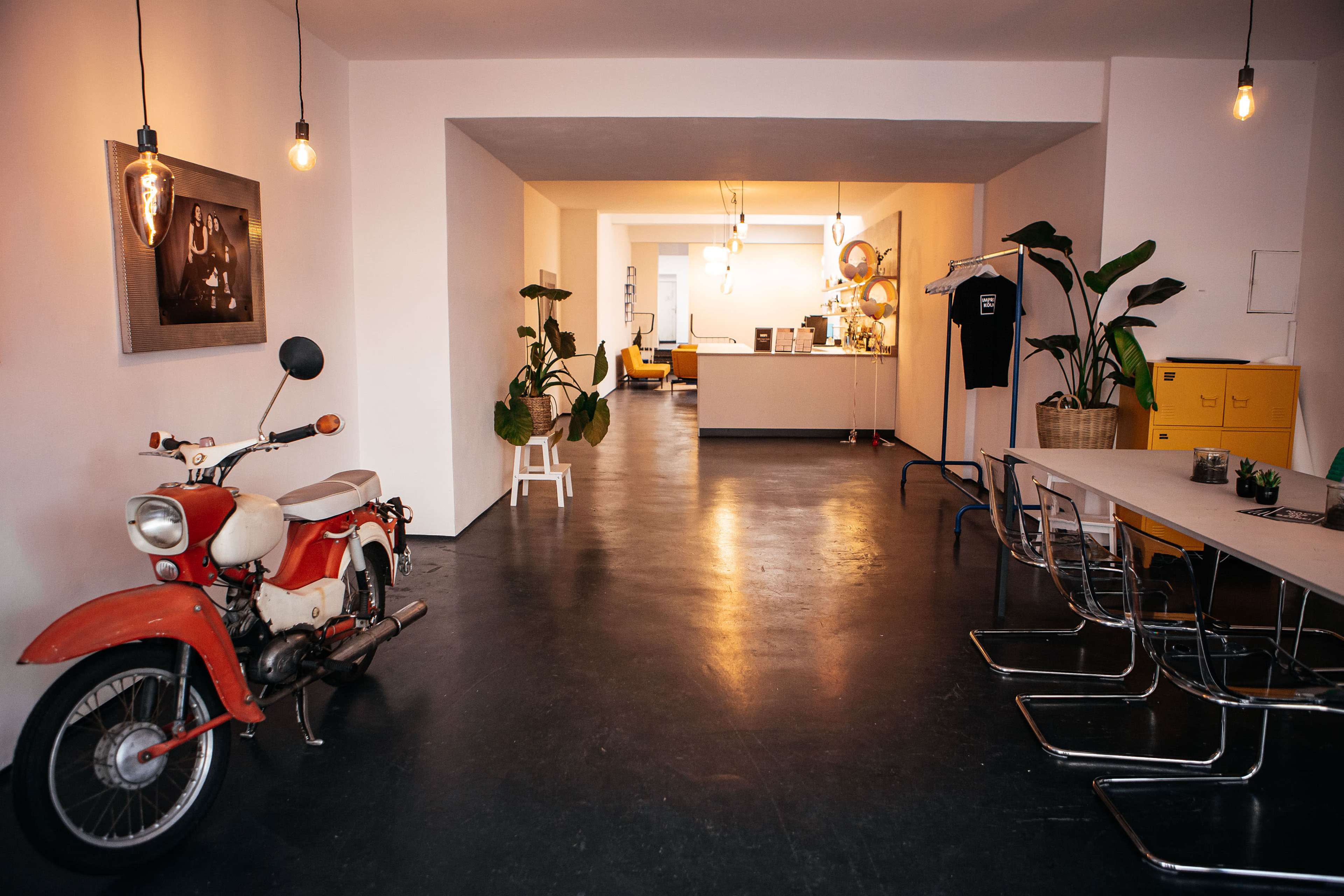 A vintage red and white motorcycle is positioned beside a modern table in a spacious, minimalist room with plants and a reception area visible in the background.