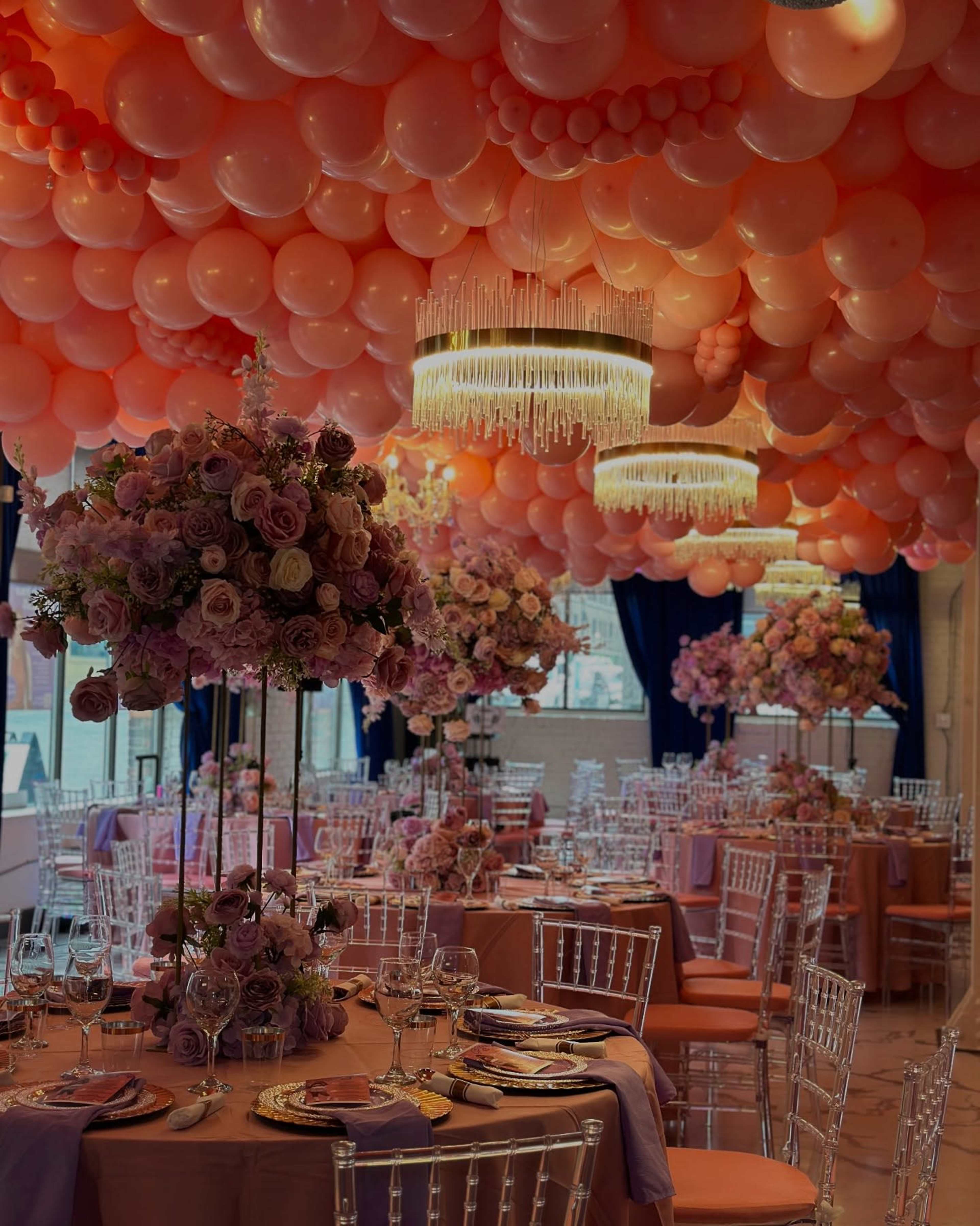 A banquet hall is decorated with a ceiling of orange balloons and tables adorned with floral centerpieces and elegant table settings.