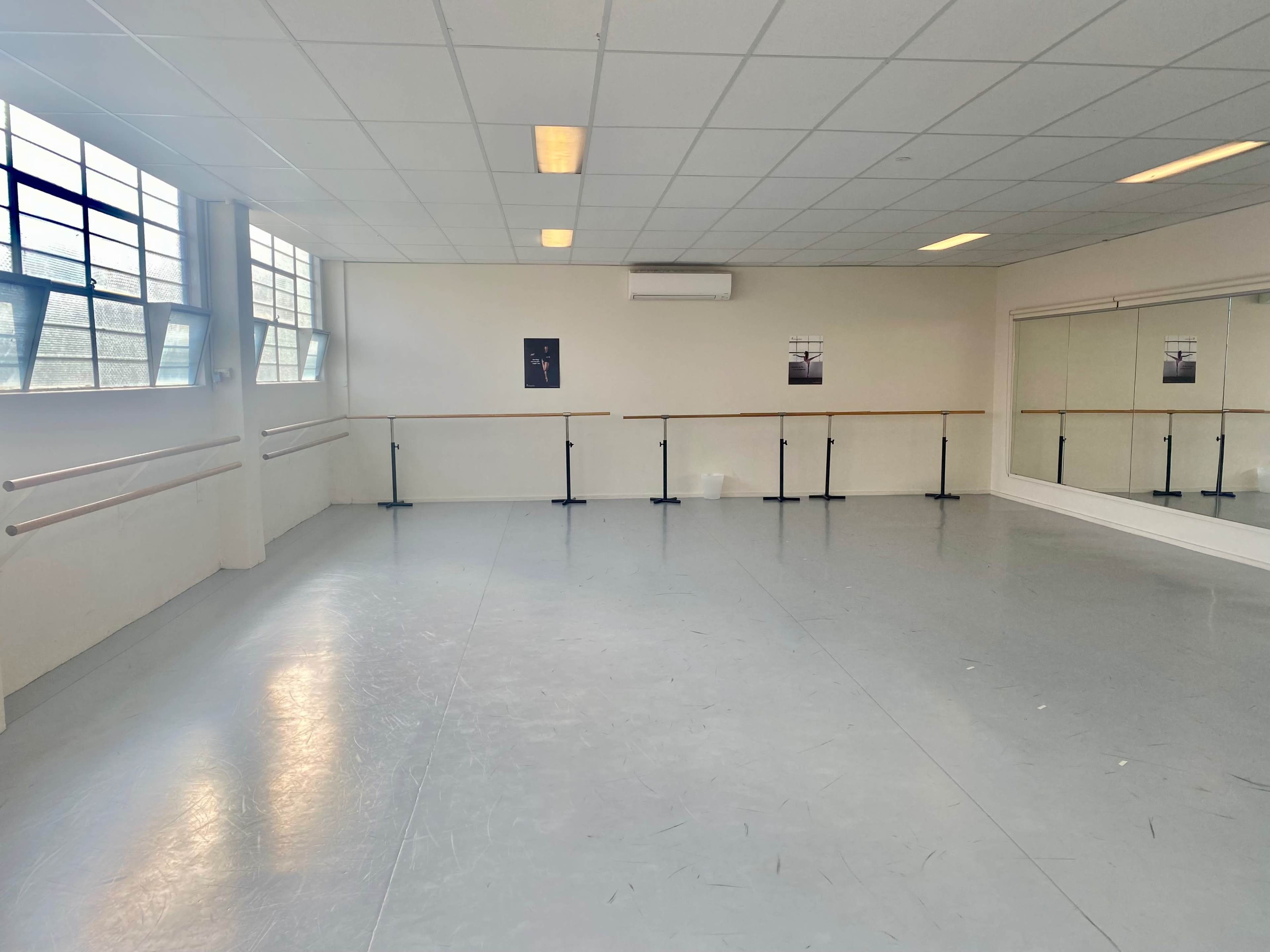A dance studio with a mirrored wall, wooden ballet barres, and large windows.