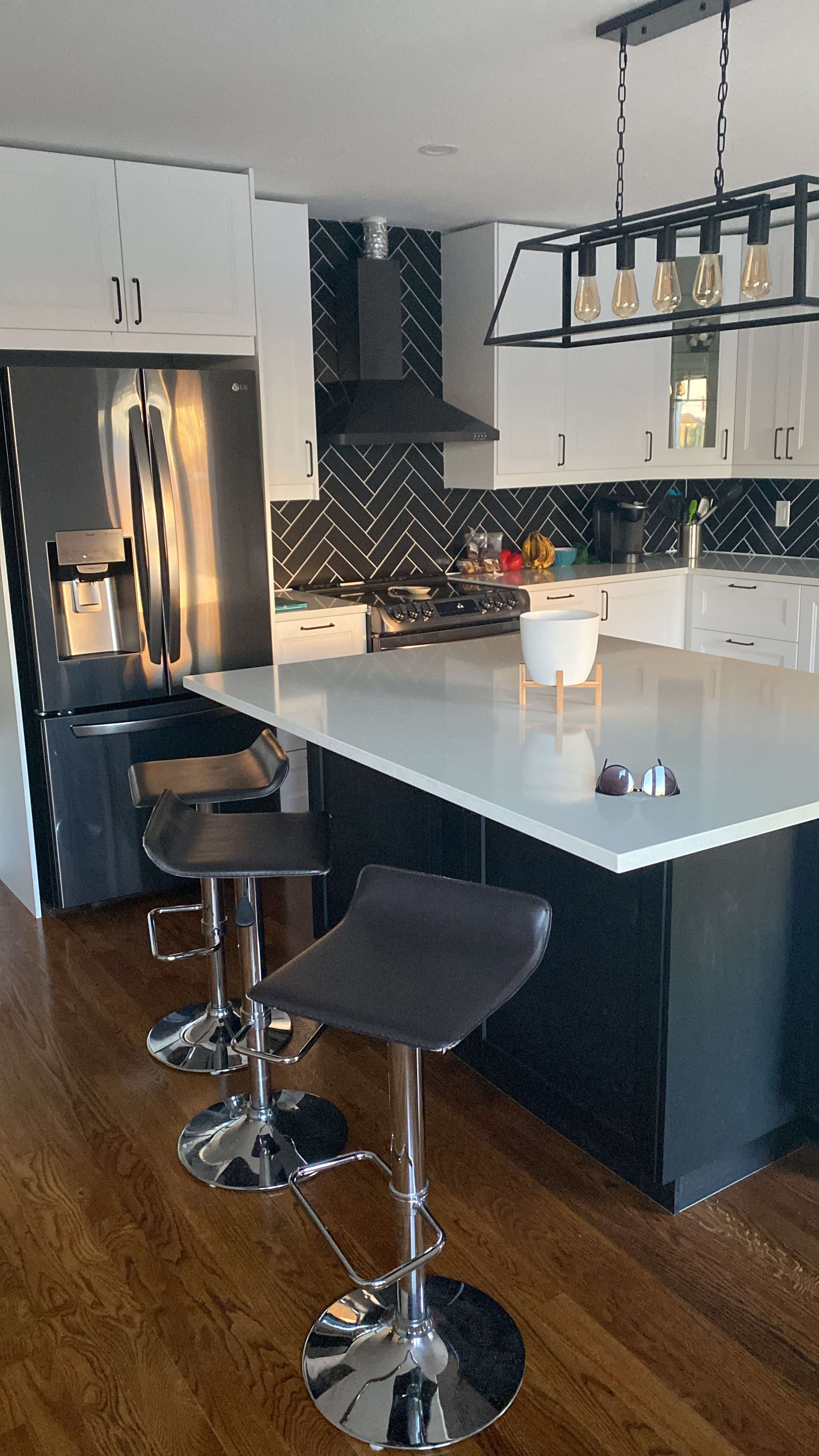 The image shows a modern kitchen featuring a black and white color scheme, an island with three bar stools, and a variety of fruits displayed on the countertop.