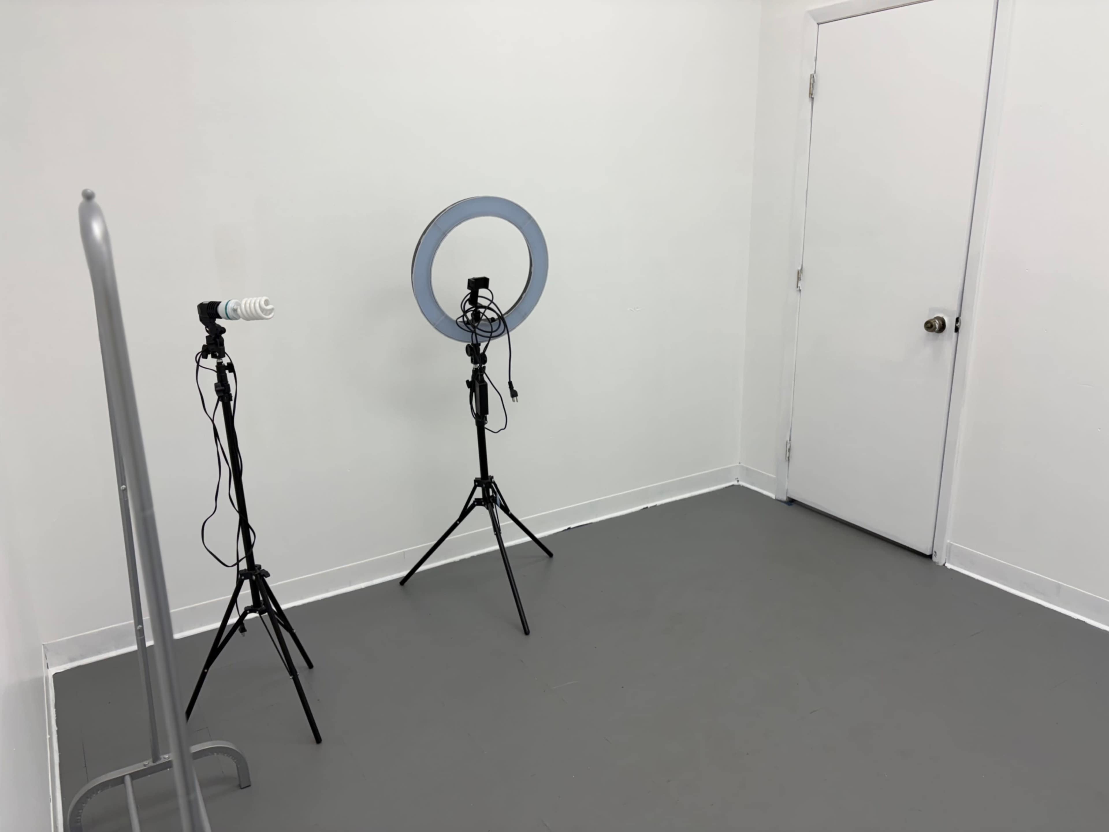 The image shows a small, empty room with two tall tripods supporting cameras and a ring light, along with a closed white door.