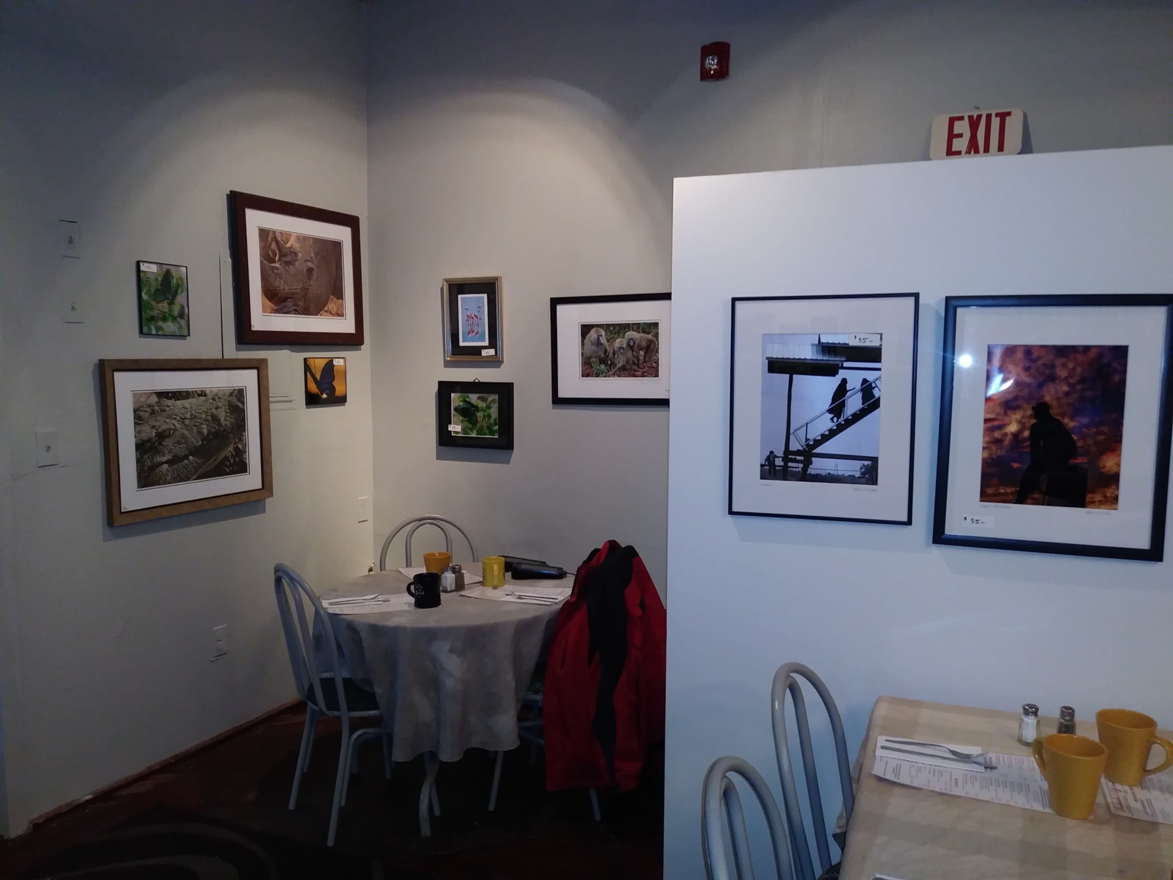 The image shows a corner of a café featuring a small round table with two chairs, surrounded by framed photographs on the walls, and an exit sign above.
