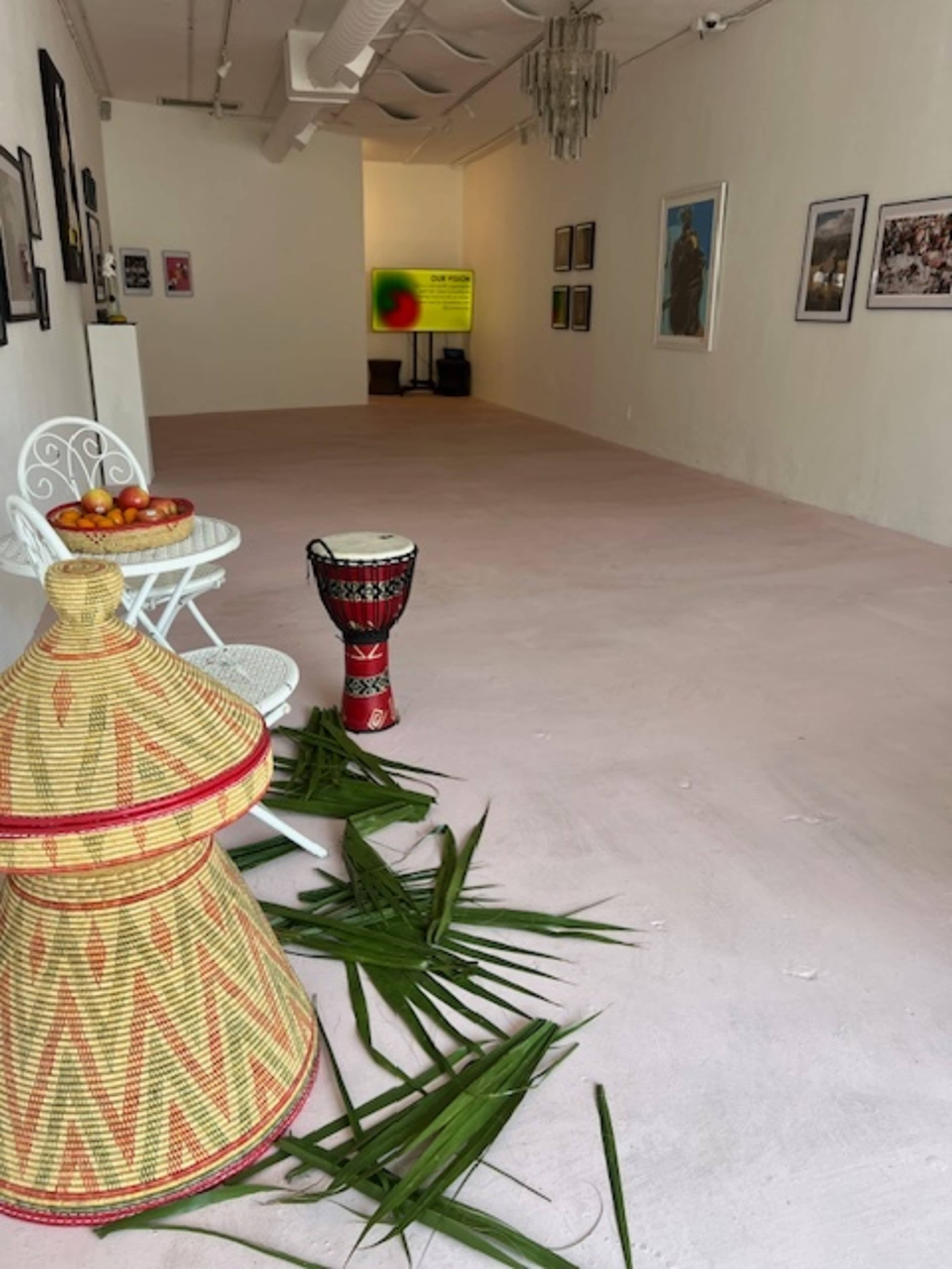 The image shows an art gallery with white walls, a light pink floor, and a display of photographs along with a television screen in the background.