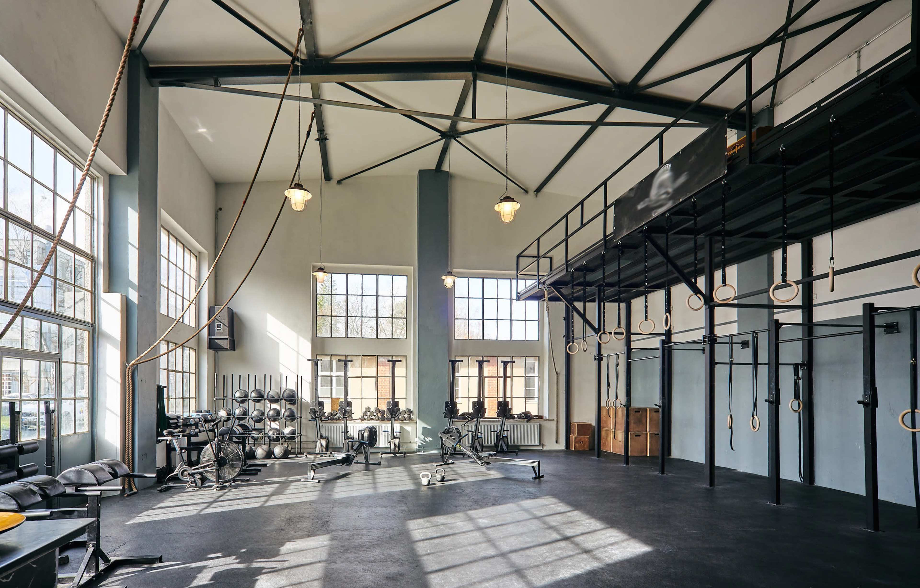 A spacious gym with large windows, fitness equipment, and a high ceiling with hanging ropes and gymnastic rings.