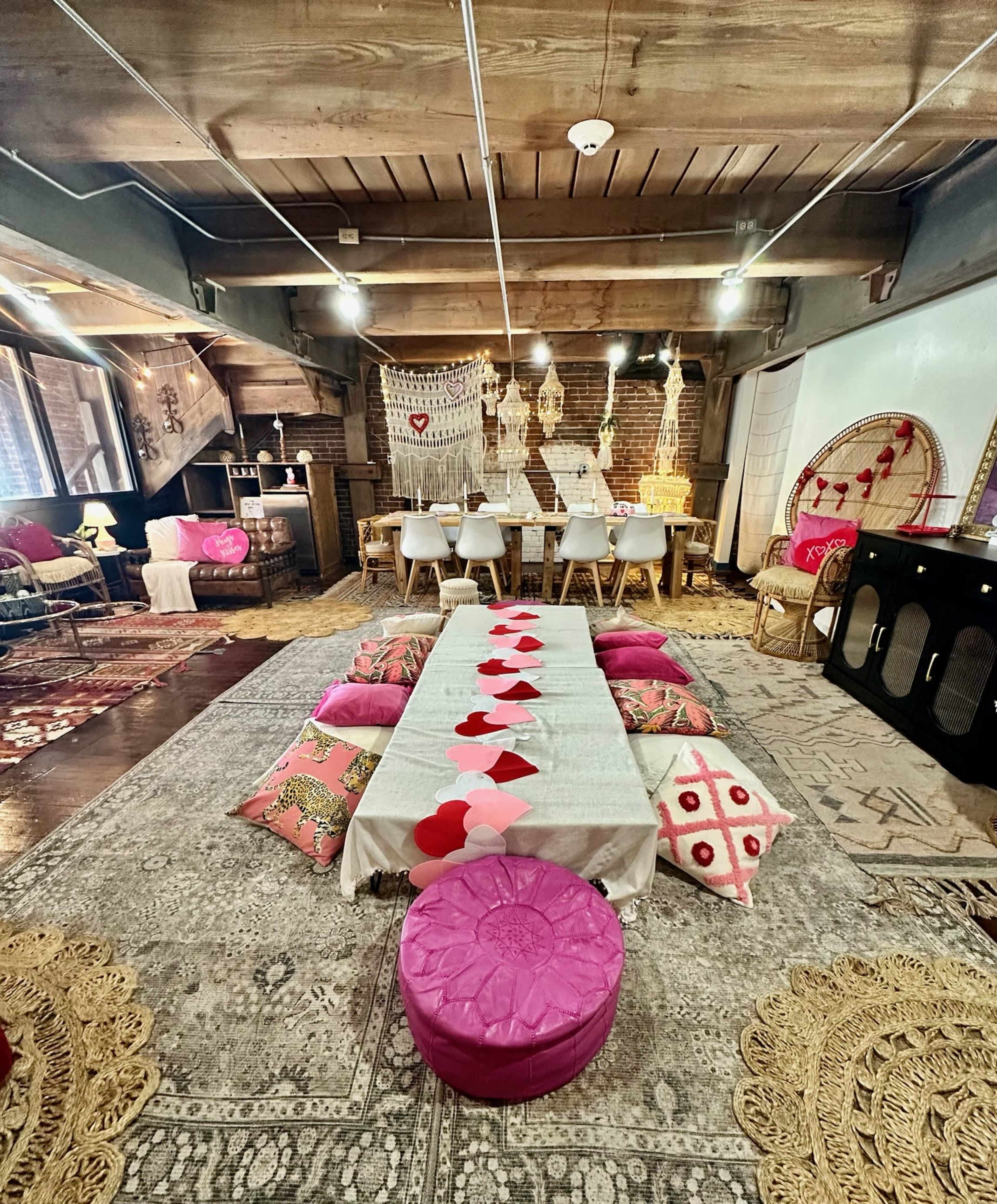 The image shows a cozy, decorated room with a long table laid out with heart-shaped decorations, surrounded by cushions and rugs, creating a warm atmosphere for gatherings.