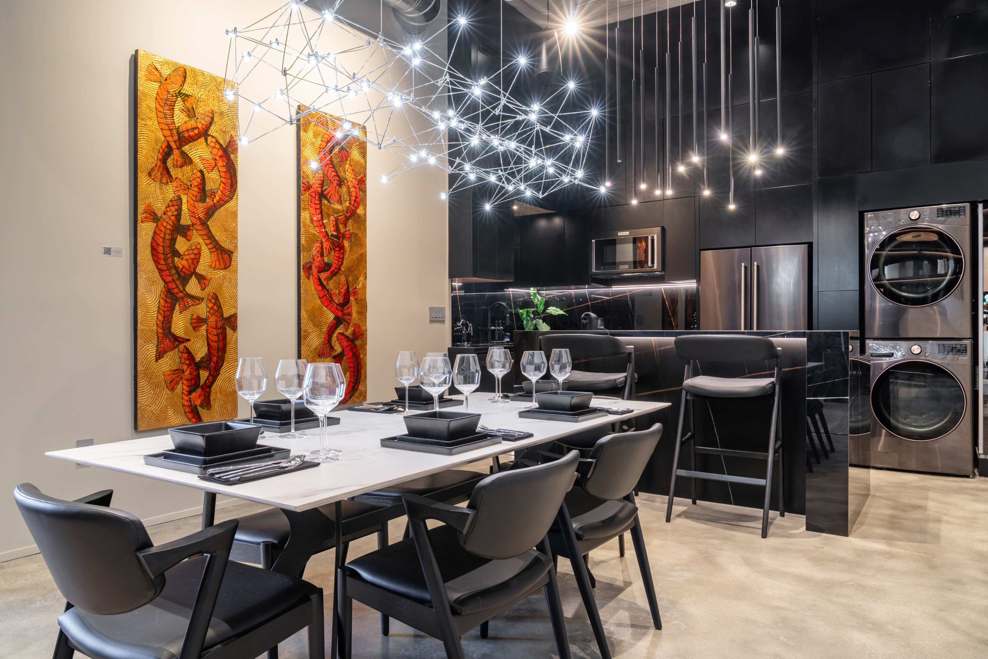 A modern dining area features a sleek table set with black dining ware and glassware, illuminated by a striking geometric light fixture and flanked by colorful artworks on the walls.