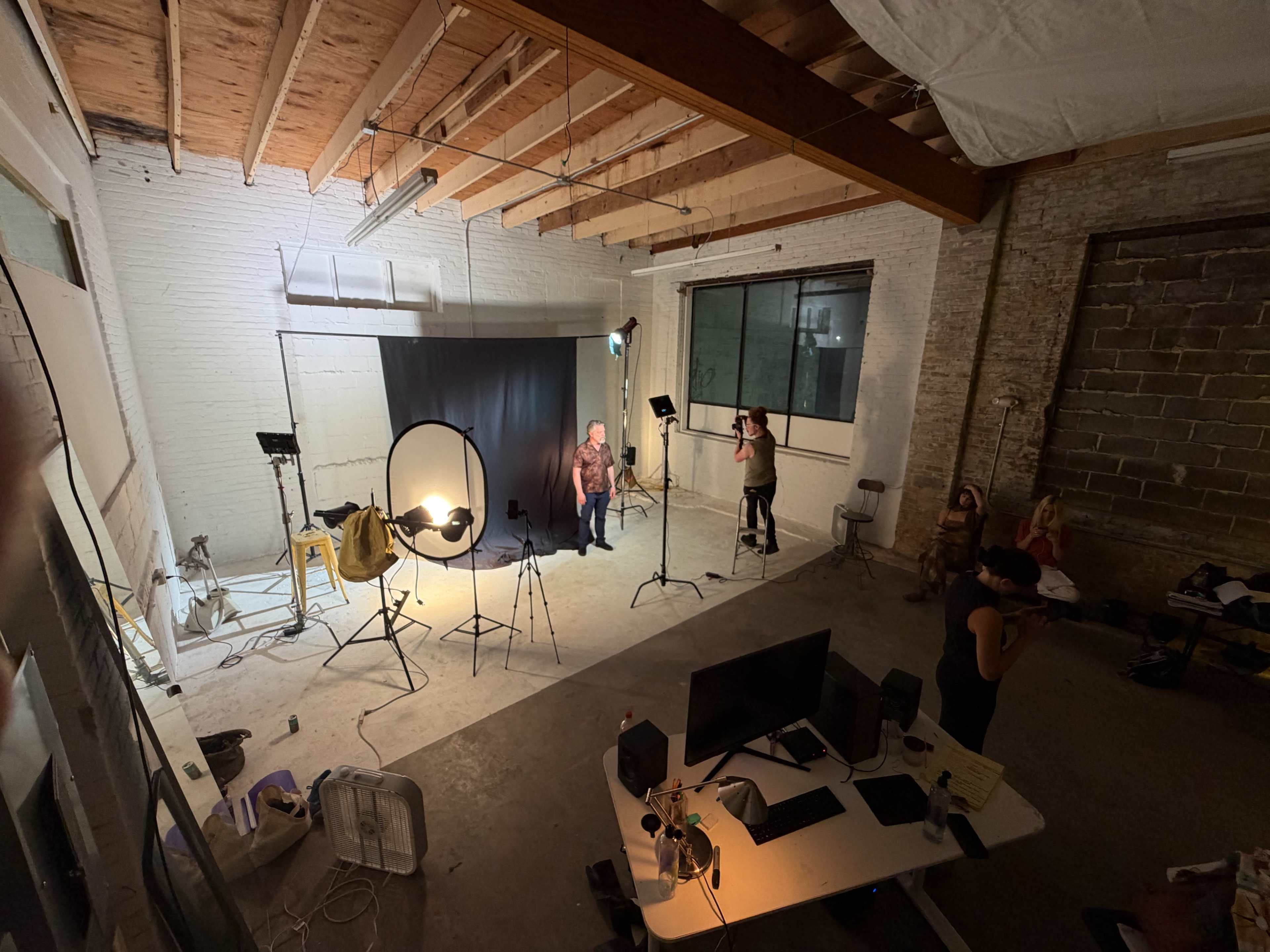 A photography studio setup features two subjects posing in front of a backdrop, surrounded by various lighting equipment and props.