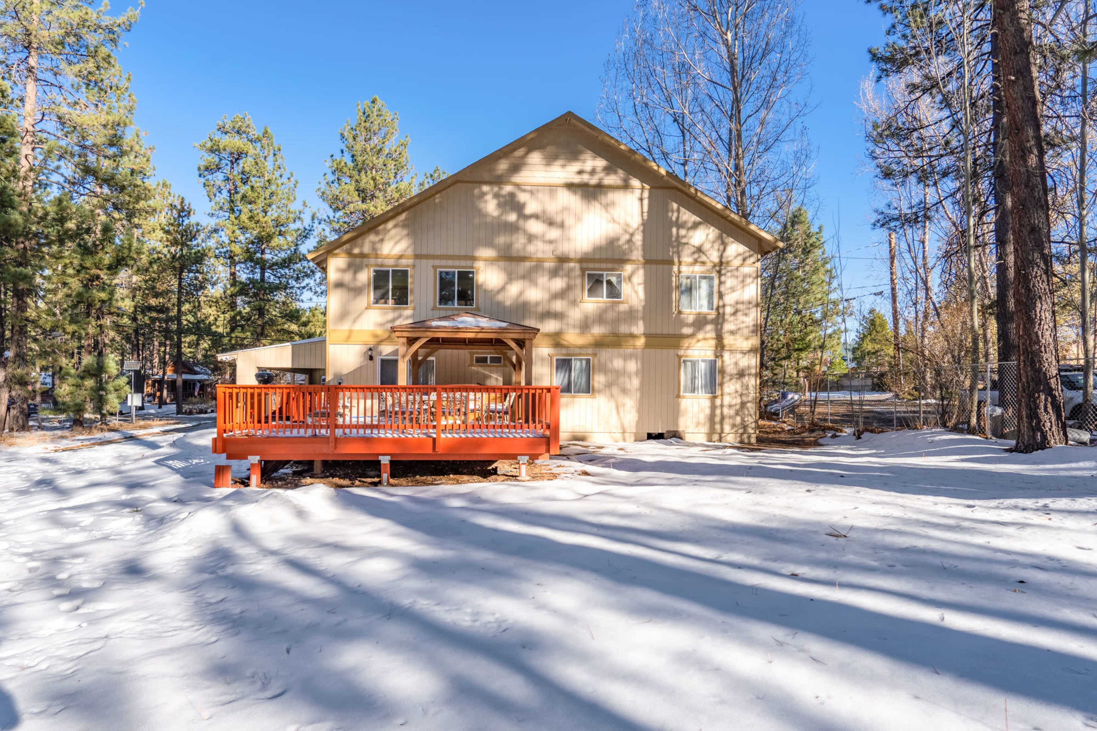 A two-story house with a wooden deck stands in a snowy landscape surrounded by pine trees.