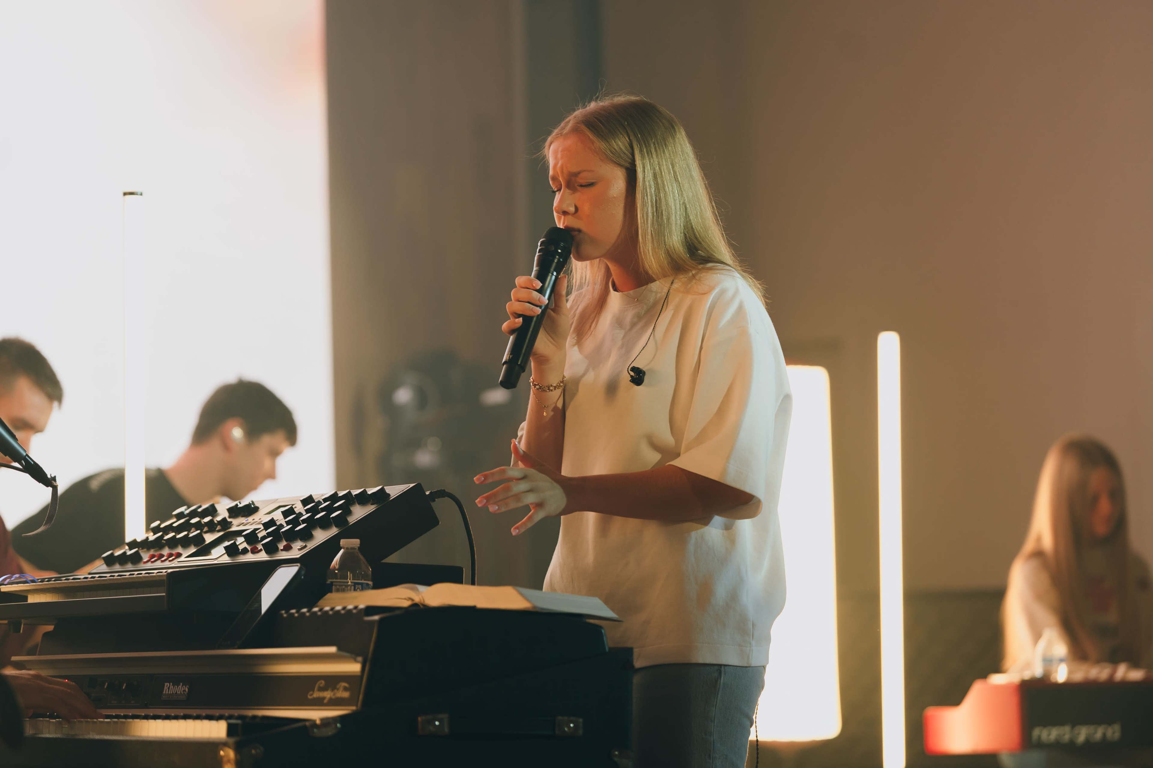 A young woman sings into a microphone while standing in front of a keyboard during a live music performance.