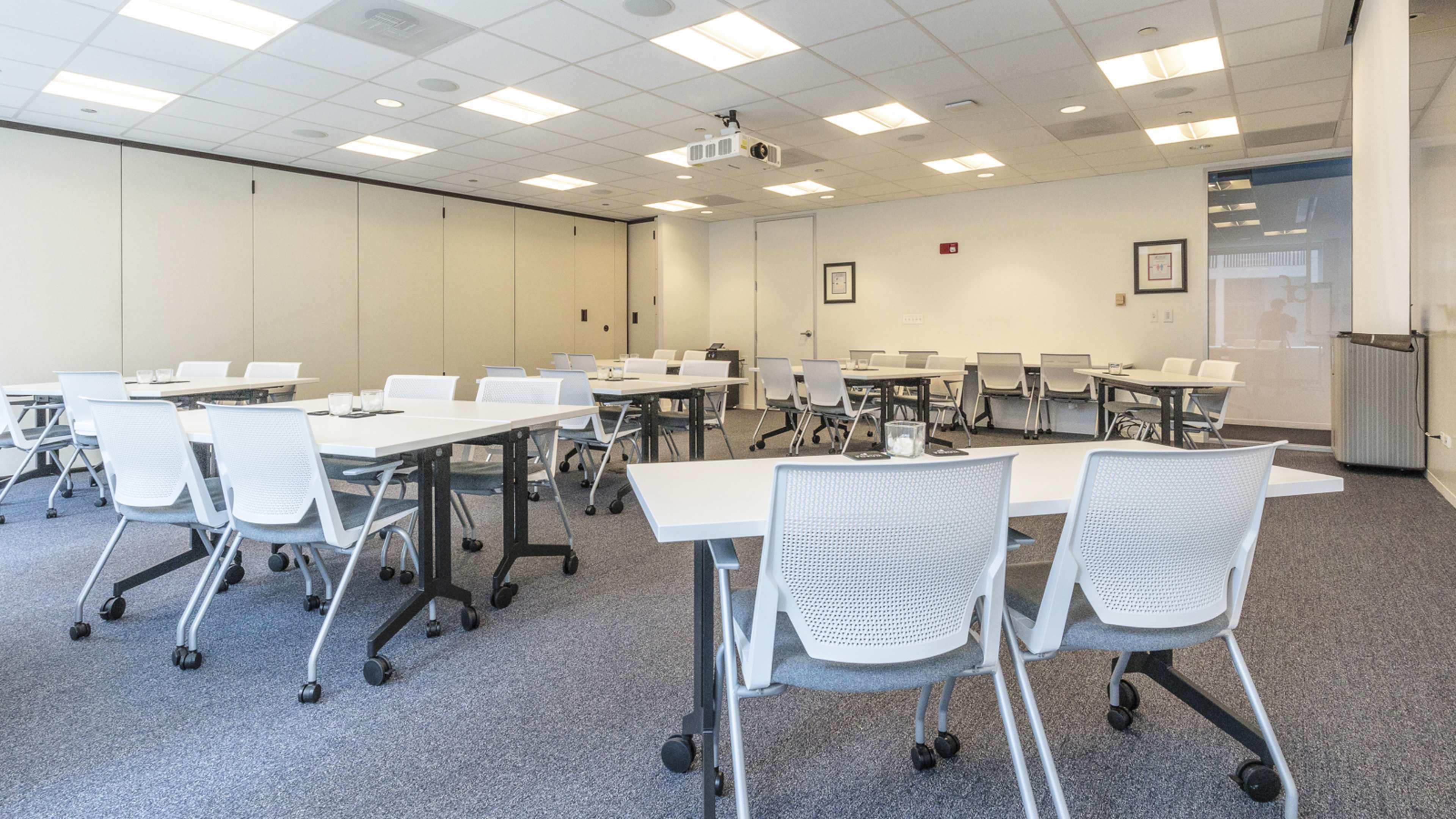 A meeting room with several white tables and chairs arranged for a presentation, equipped with a projector and a light-colored carpet.