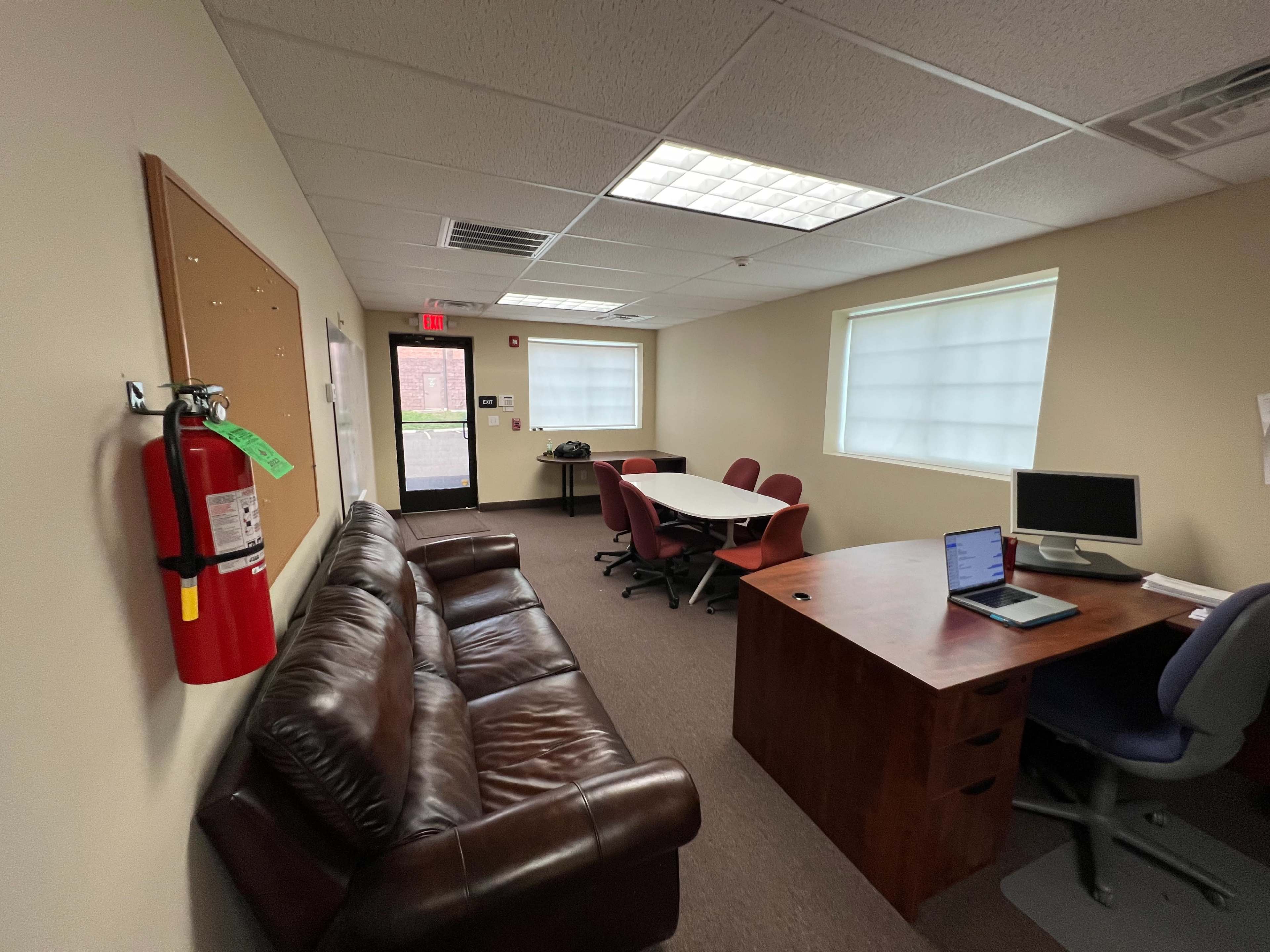 An office space featuring a dark brown leather couch, a wooden desk with a laptop, several chairs, a white table, and a fire extinguisher on the wall.