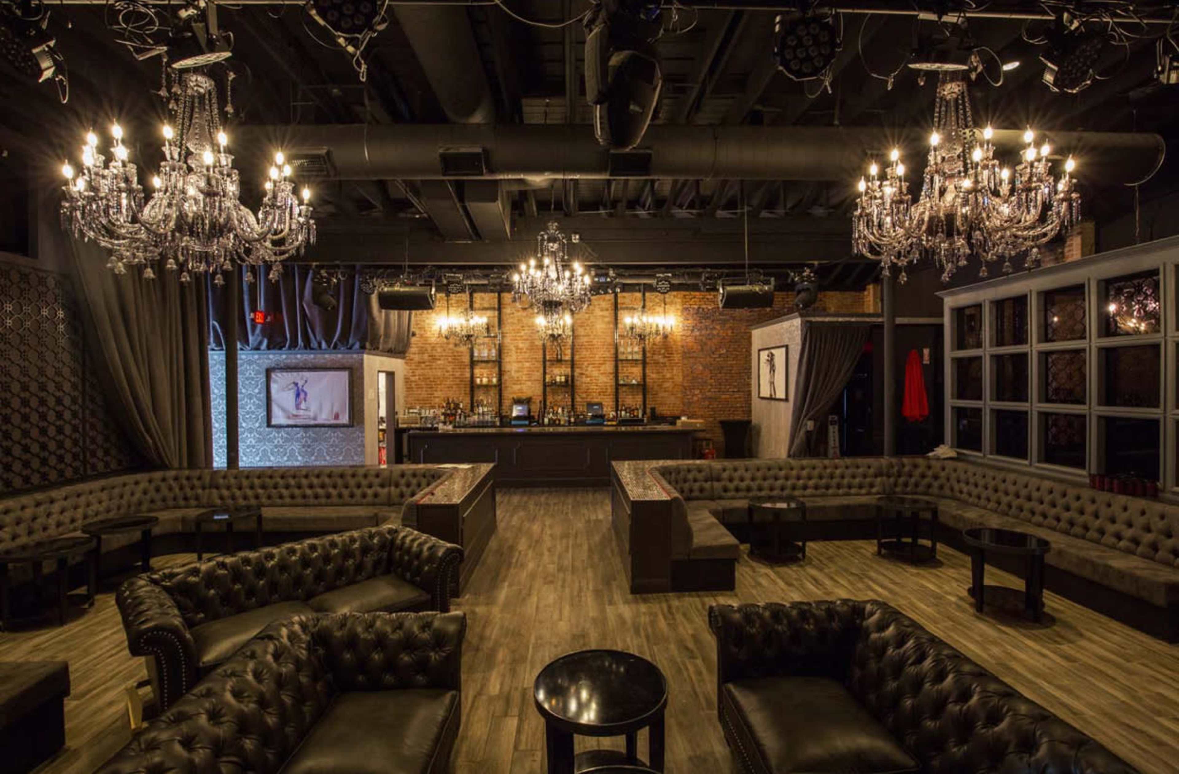 The image shows an upscale lounge with dark leather seating, ornate chandeliers, and a well-stocked bar in the background against a brick wall.