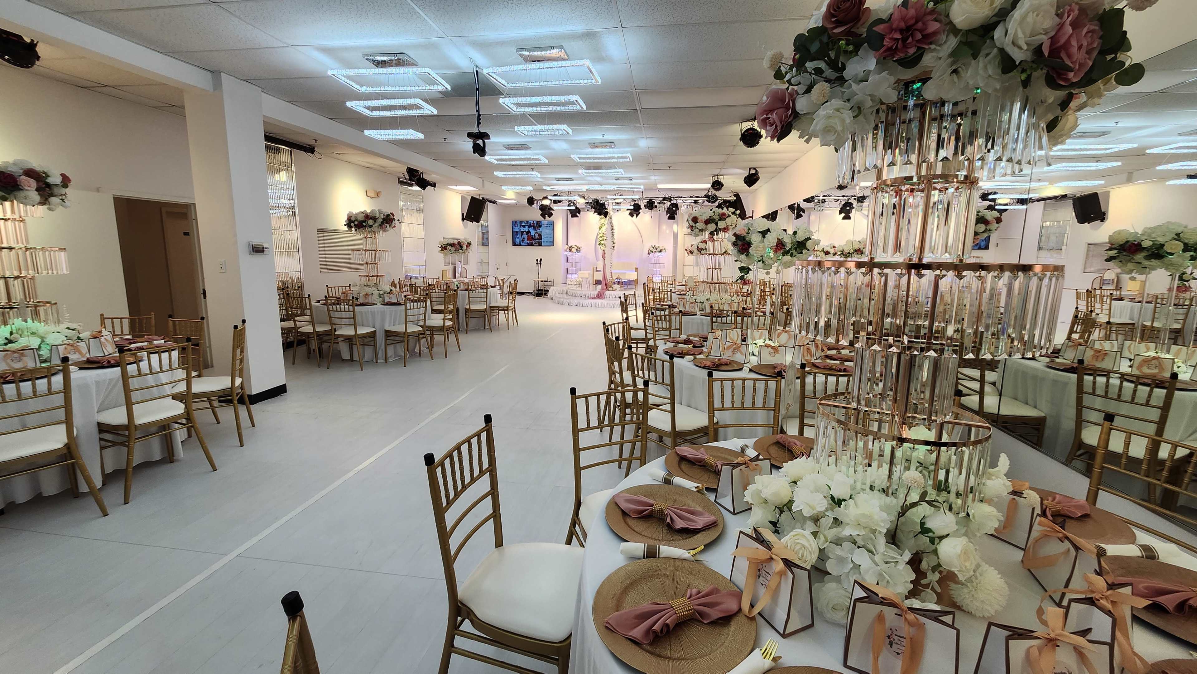 The image shows a banquet hall set up for an event, with elegantly arranged tables featuring floral centerpieces and golden chairs.