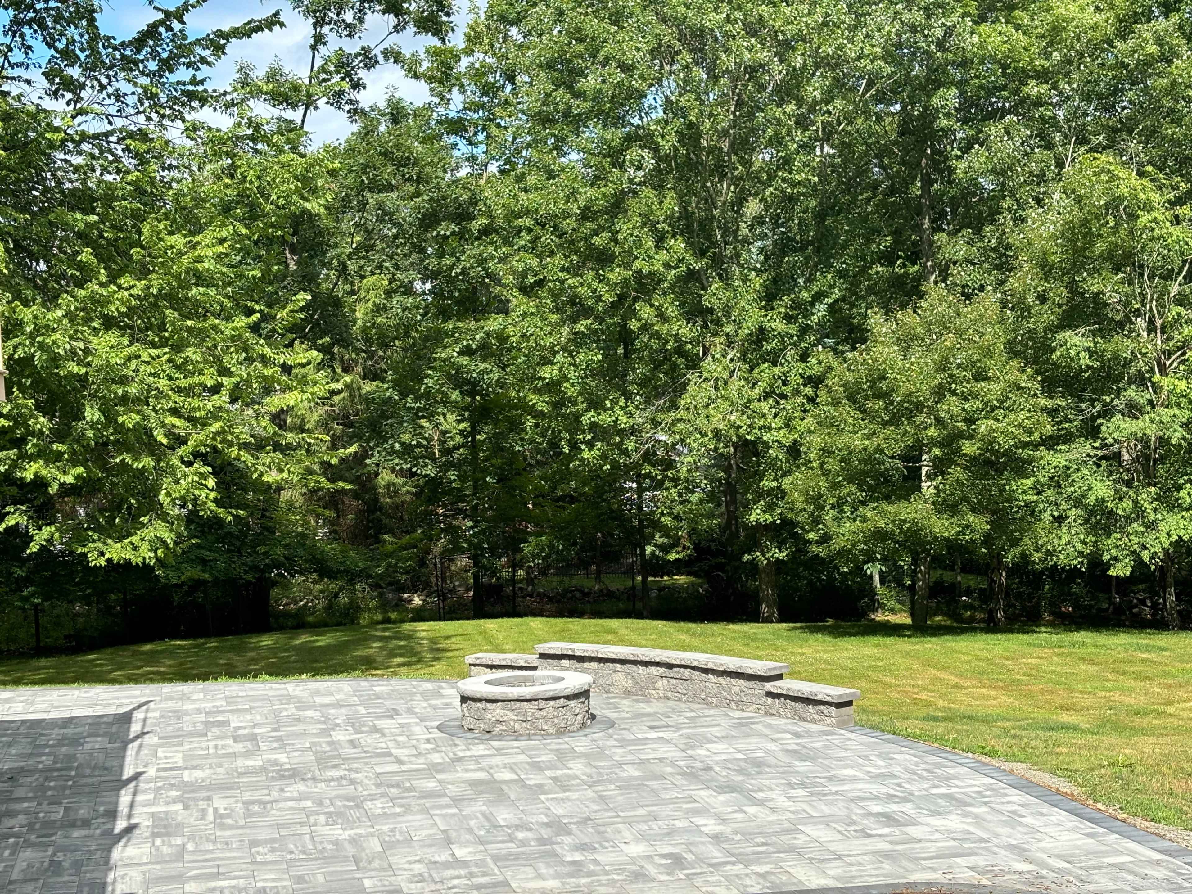 A stone patio with a circular seating area surrounded by lush green trees and a grassy lawn.