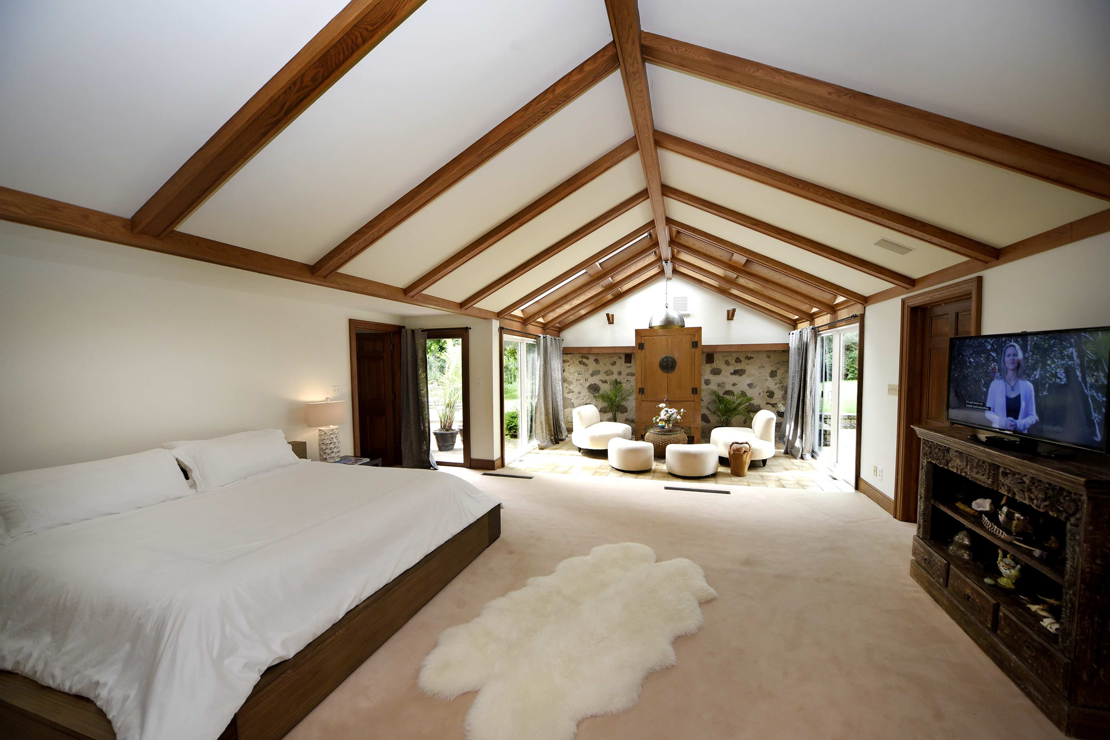 A spacious bedroom with a king-sized bed, wooden beams on the ceiling, and a seating area near large windows overlooking a garden.