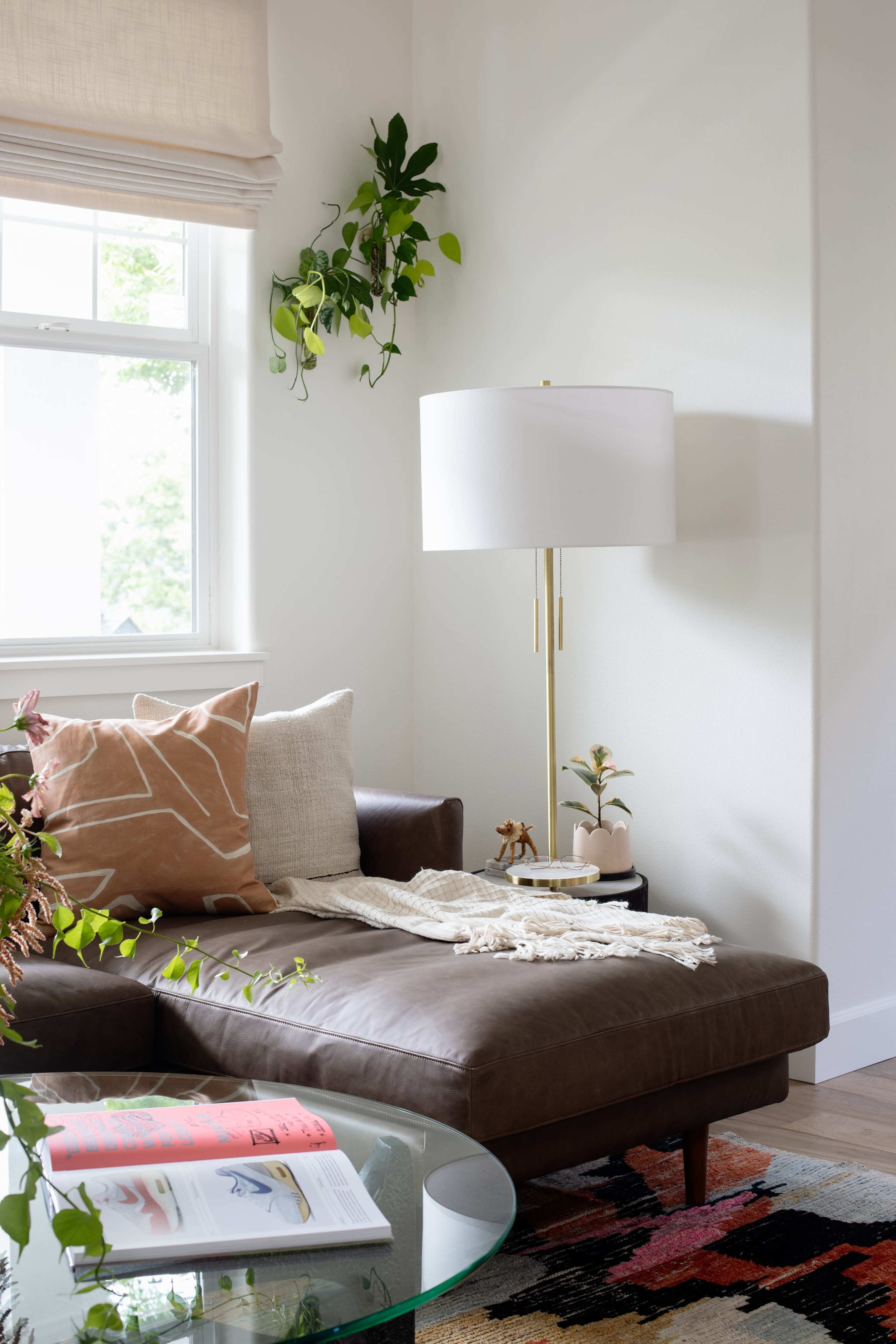 A modern living room features a brown leather sofa adorned with decorative pillows, a large floor lamp, a glass coffee table, and greenery by the window.