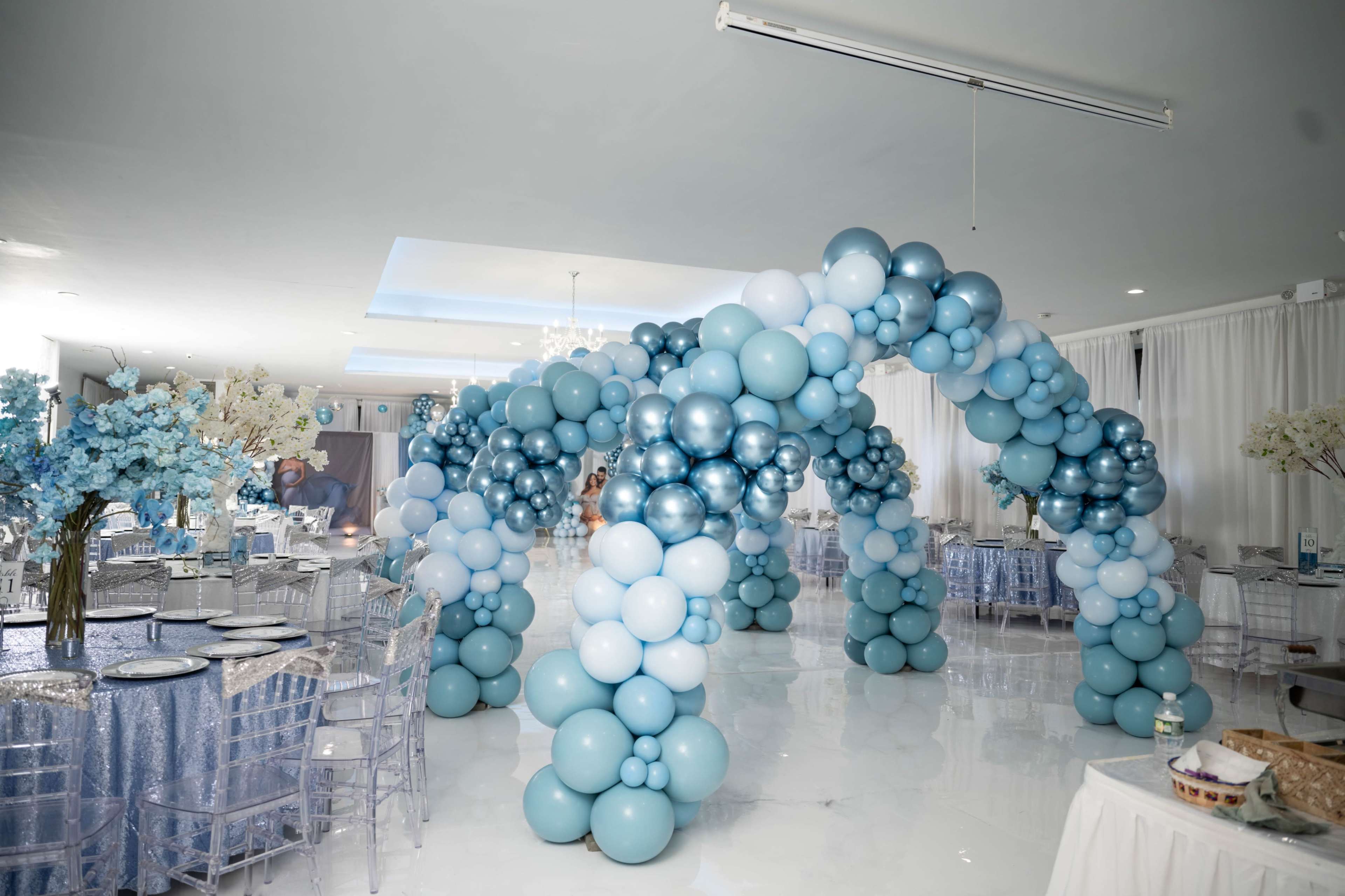 A large indoor event space features an archway made of blue and silver balloons, surrounded by elegantly set tables with blue tablecloths and clear chairs.