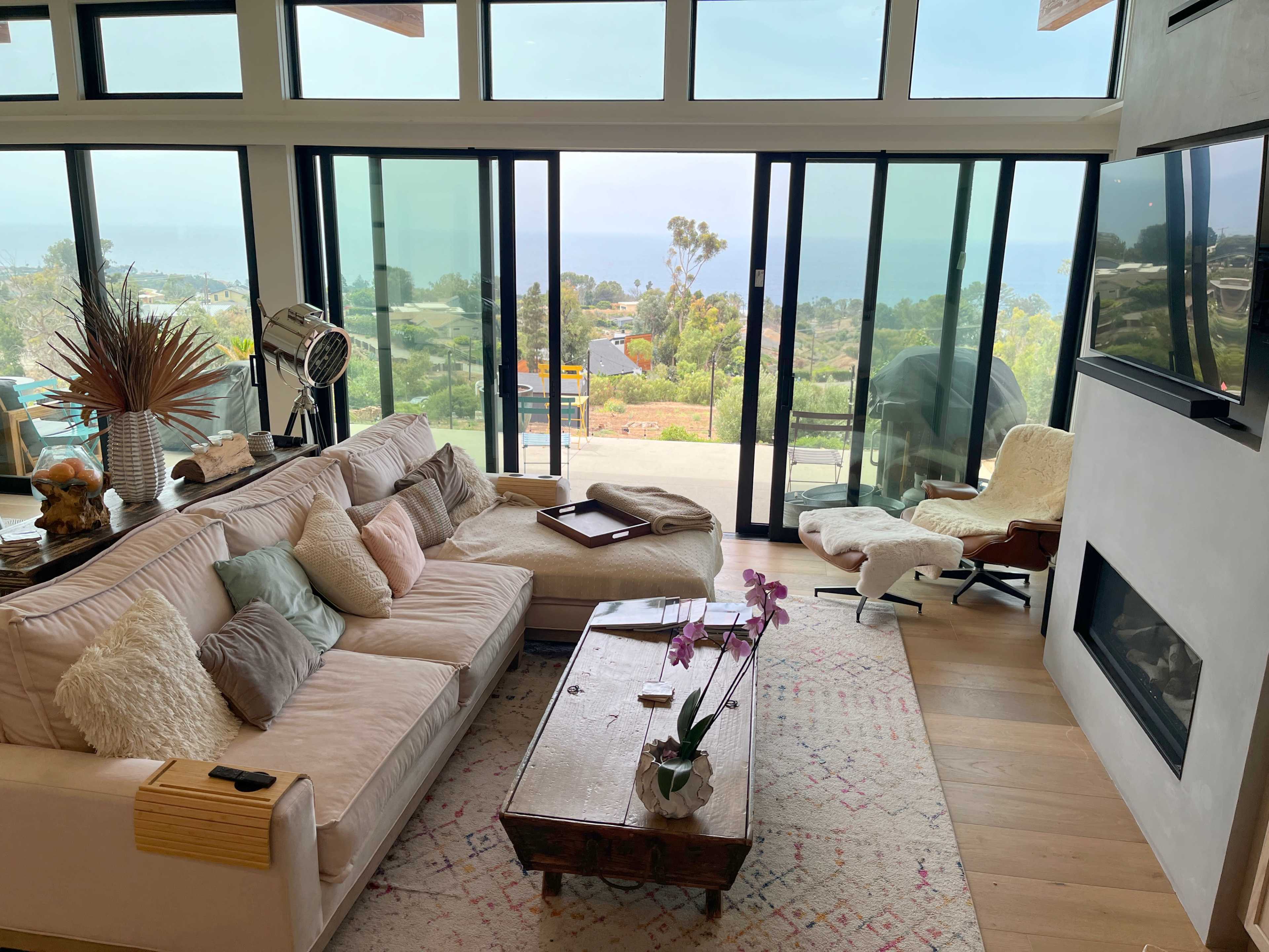 The image shows a modern living room with large windows overlooking a coastal view, featuring a sectional sofa, a wooden coffee table, and a lounge chair.