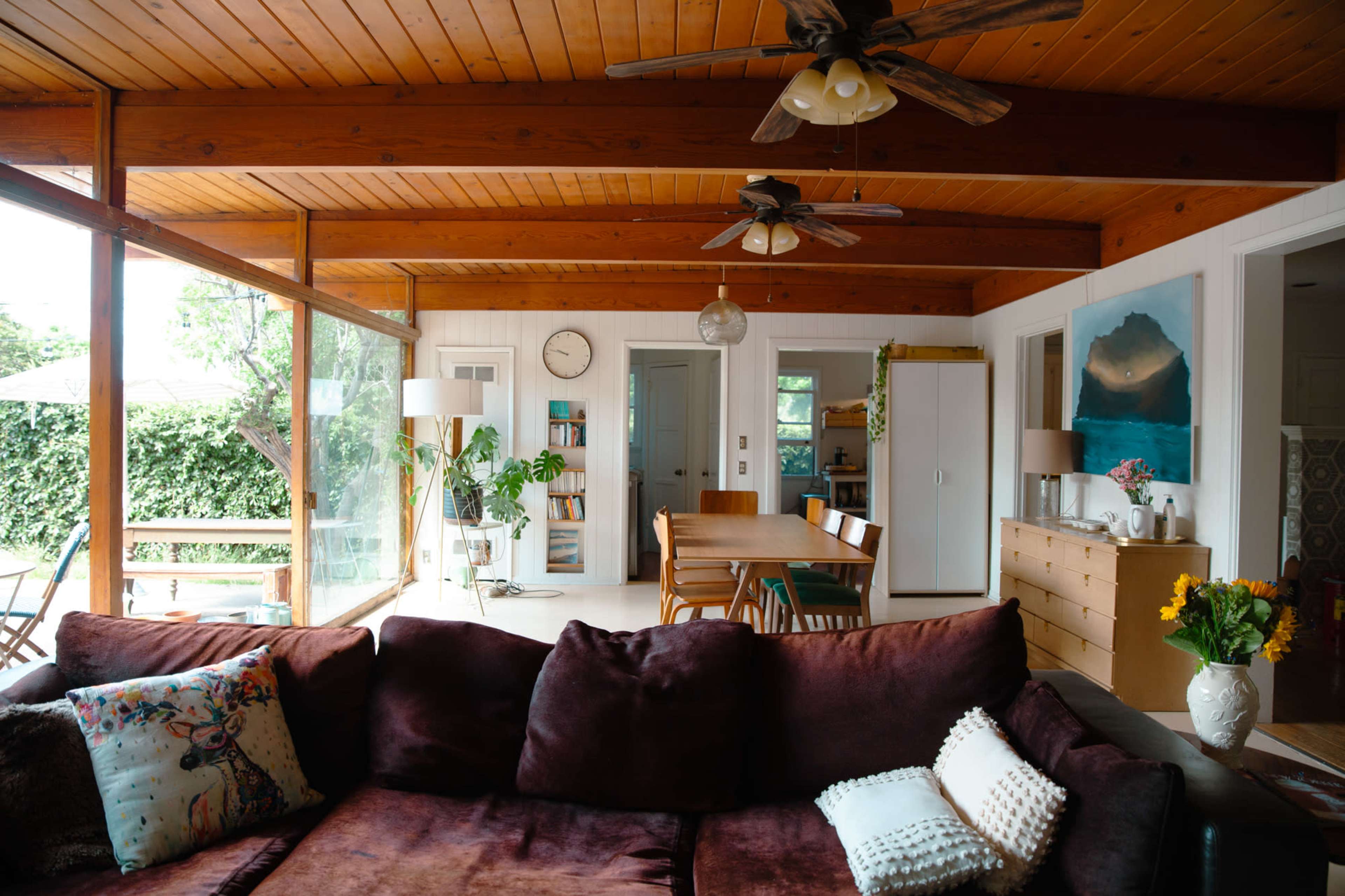 A spacious living room features a large brown sofa, wooden beams overhead, and a dining area with a table, surrounded by large windows that overlook a green outdoor space.