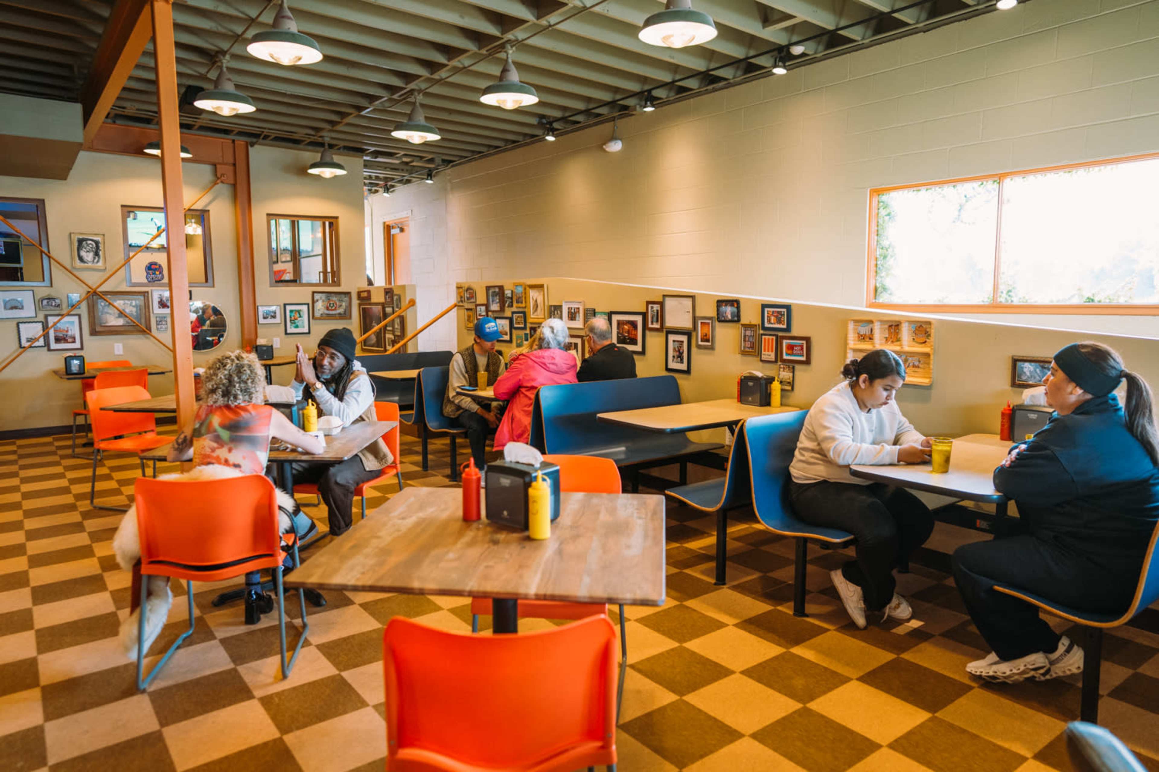 The image shows a casual dining area with several tables occupied by customers, featuring a mix of chairs and artwork on the walls.