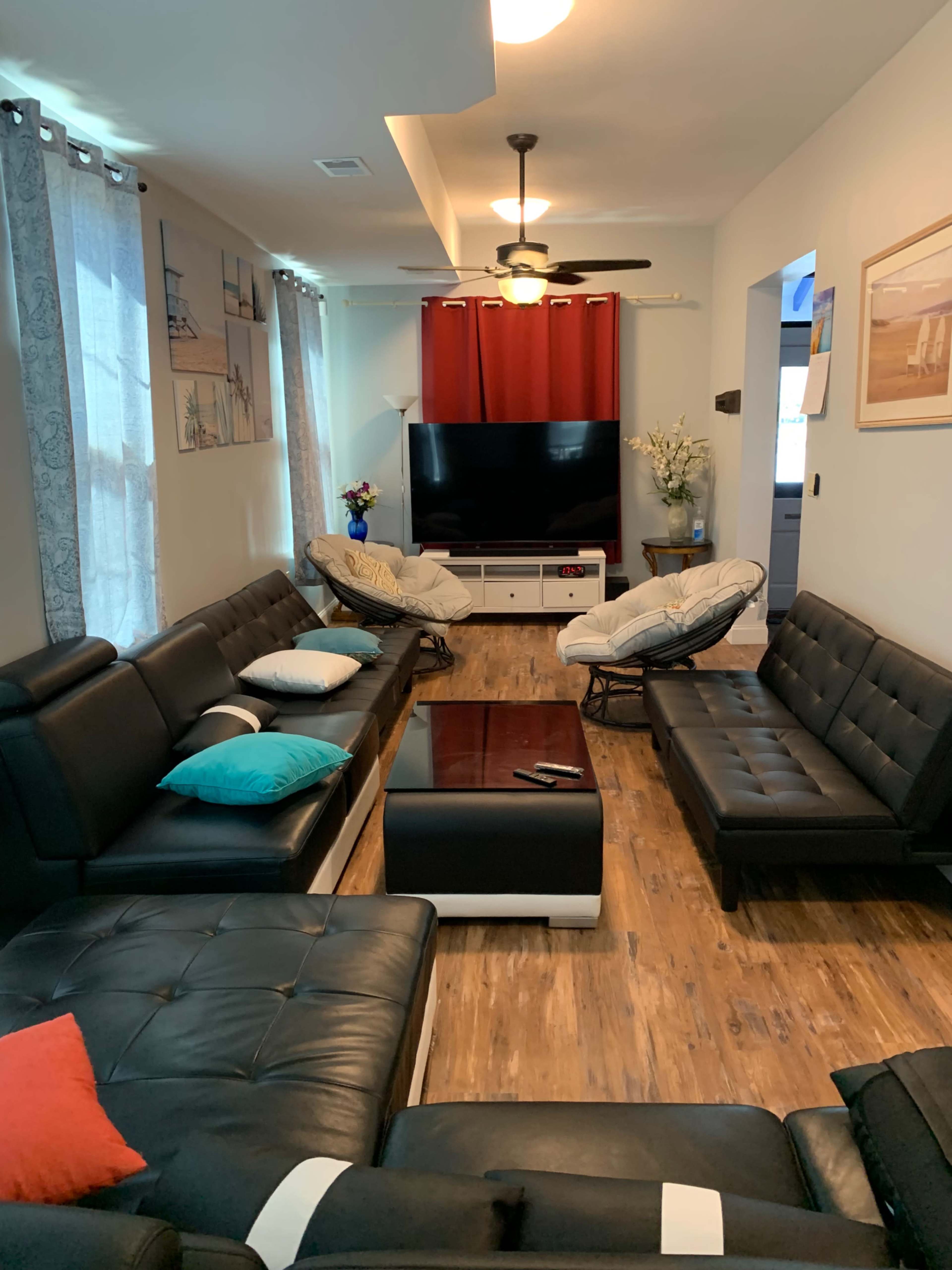 The living room features black leather sofas arranged around a central coffee table, with a large television and decorative plants in the background.
