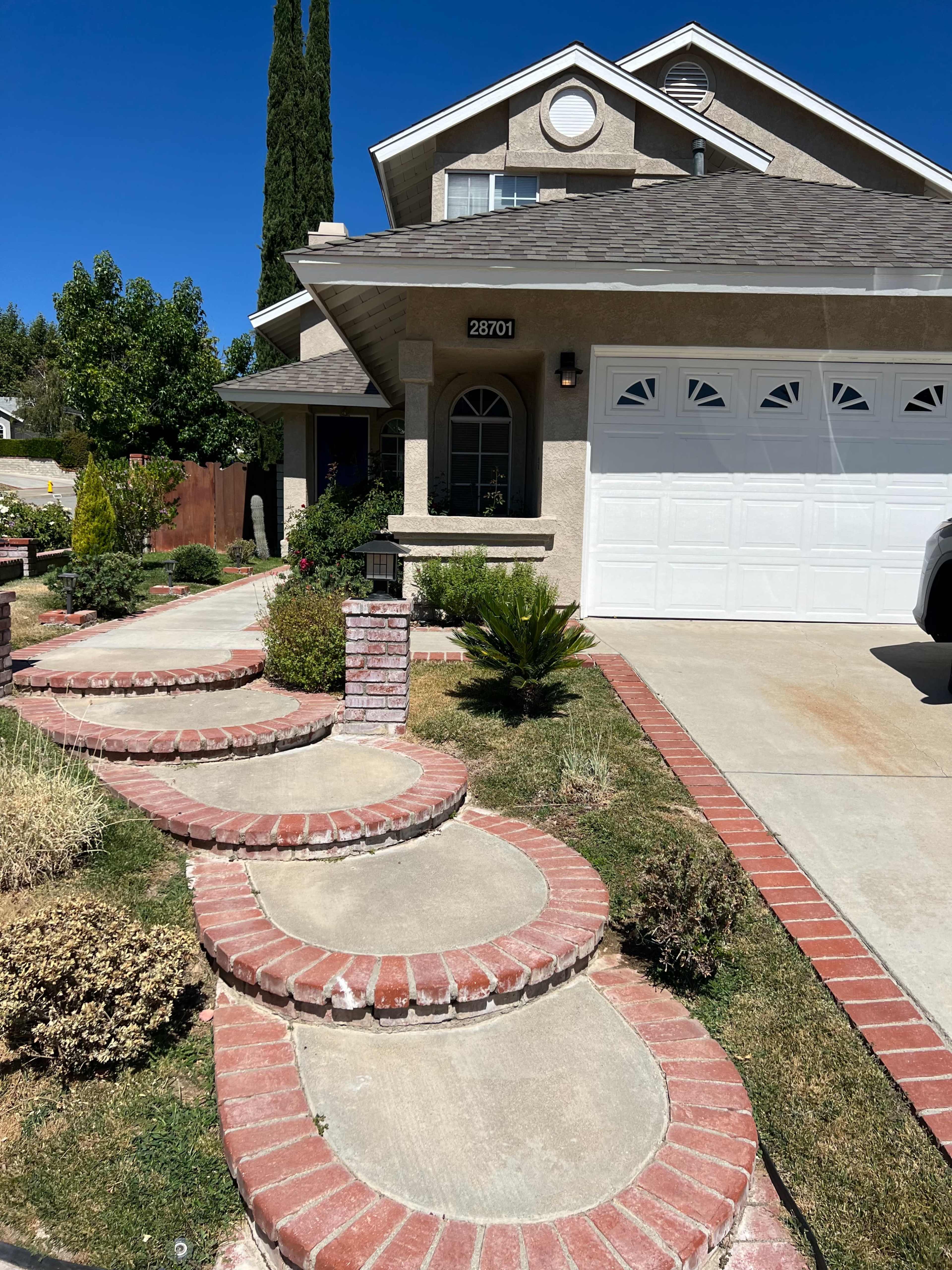 A residential front yard featuring a winding brick pathway leading to a house with a garage.