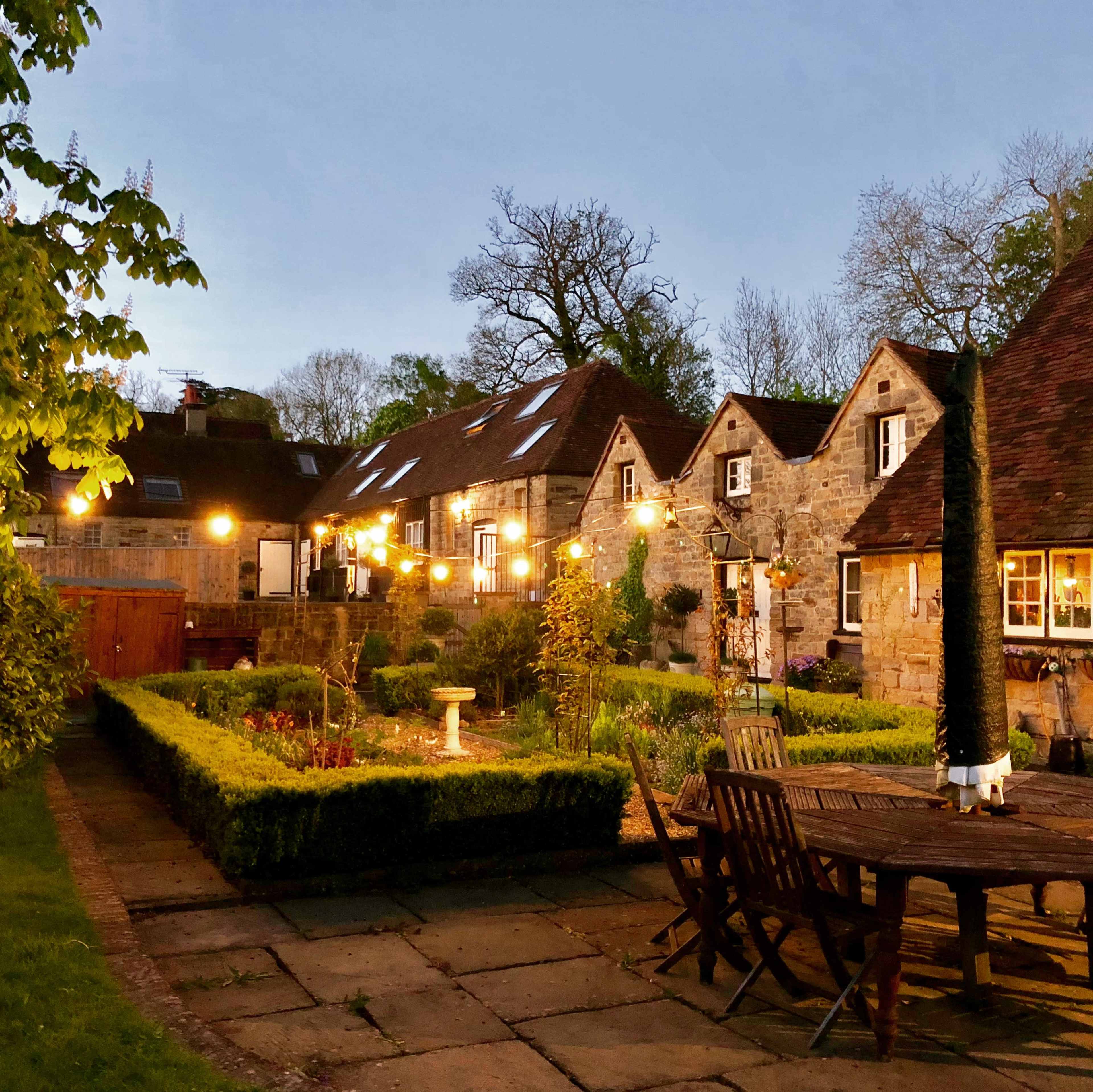 A stone cottage with a landscaped garden and string lights is illuminated in the evening twilight.
