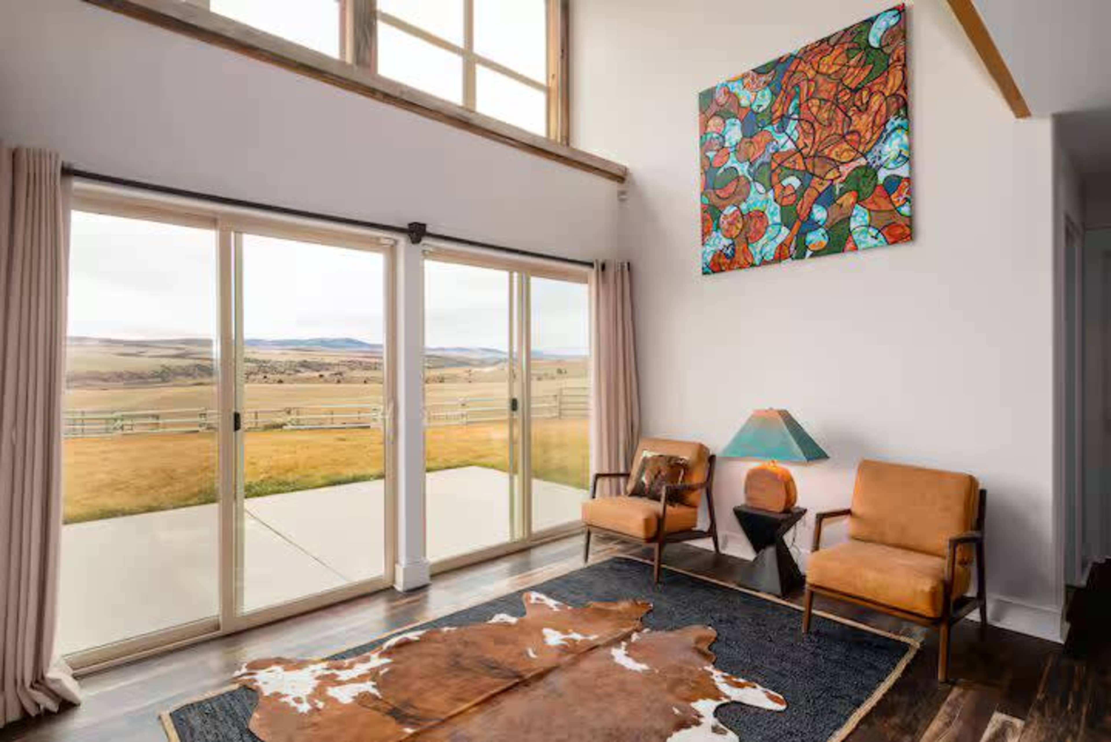 The image shows a spacious living area with large sliding glass doors that open to a view of rolling hills, furnished with two leather chairs, a decorative lamp, and a cowhide rug on the floor.