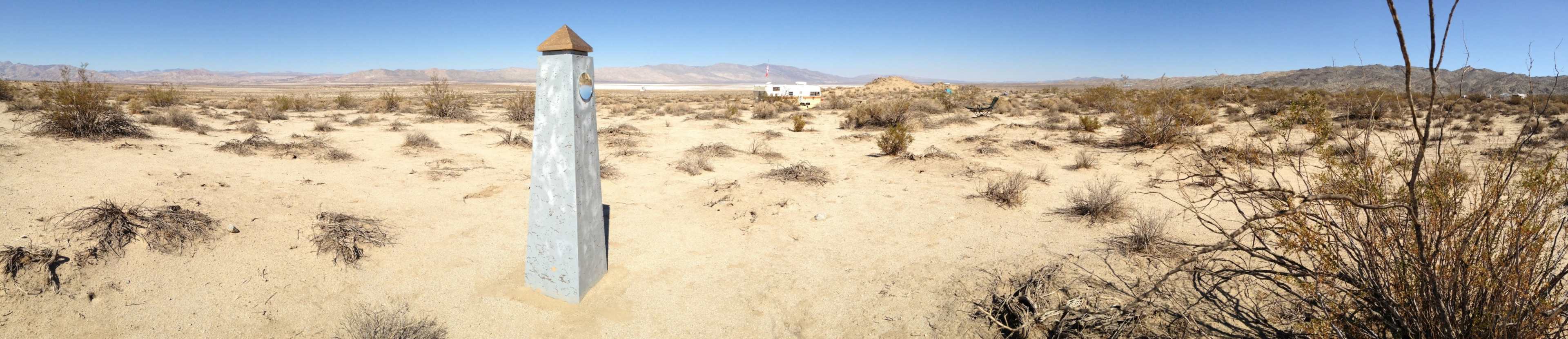 A tall, triangular stone marker stands in a dry, desert landscape surrounded by sparse vegetation and distant mountains.