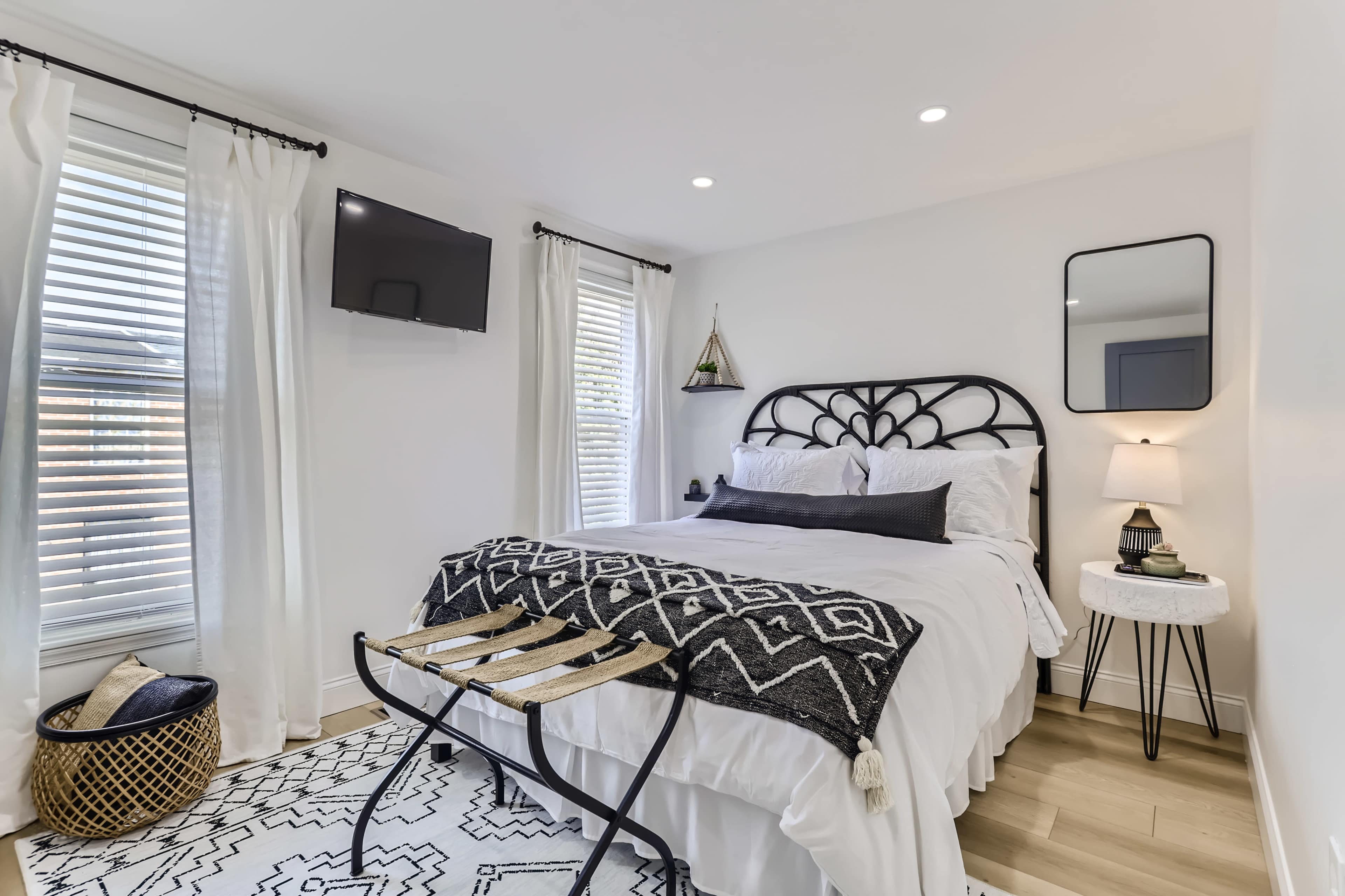 A modern bedroom features a black iron bed frame, white linens, a small flat-screen TV, and a patterned rug, with natural light streaming through two windows.